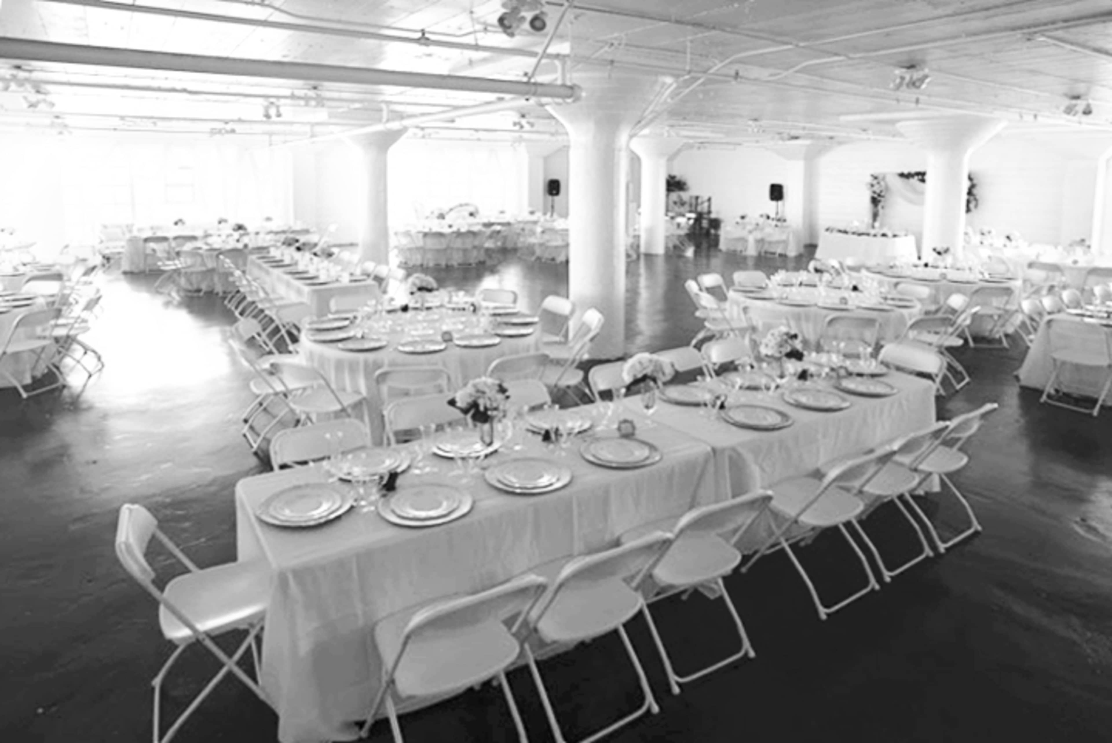 A spacious event venue features neatly arranged dining tables covered with white tablecloths and set with plates and glasses.
