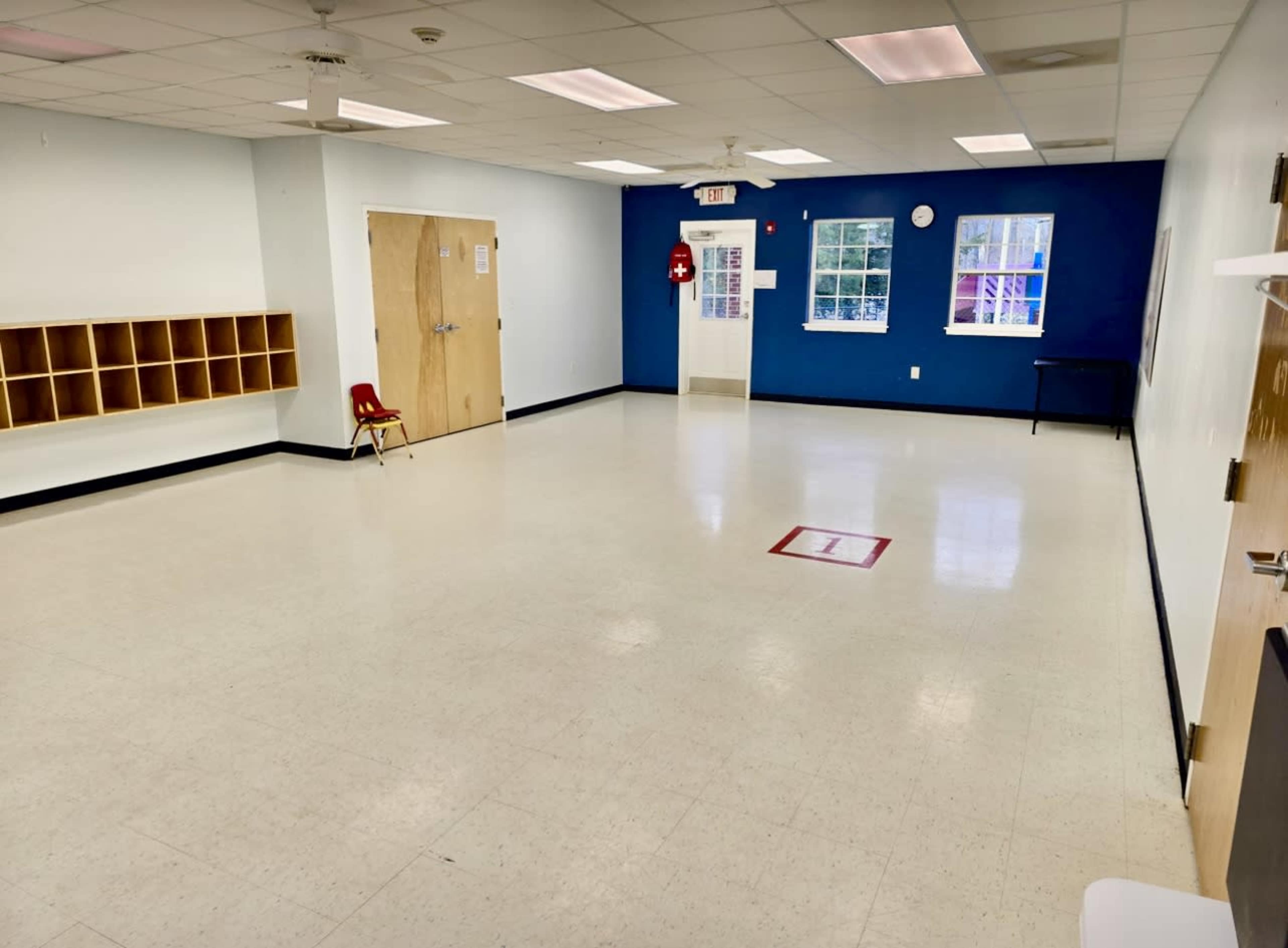 The image shows a spacious, empty classroom with a blue accent wall, a row of cubbies, a red chair, and minimal furniture.