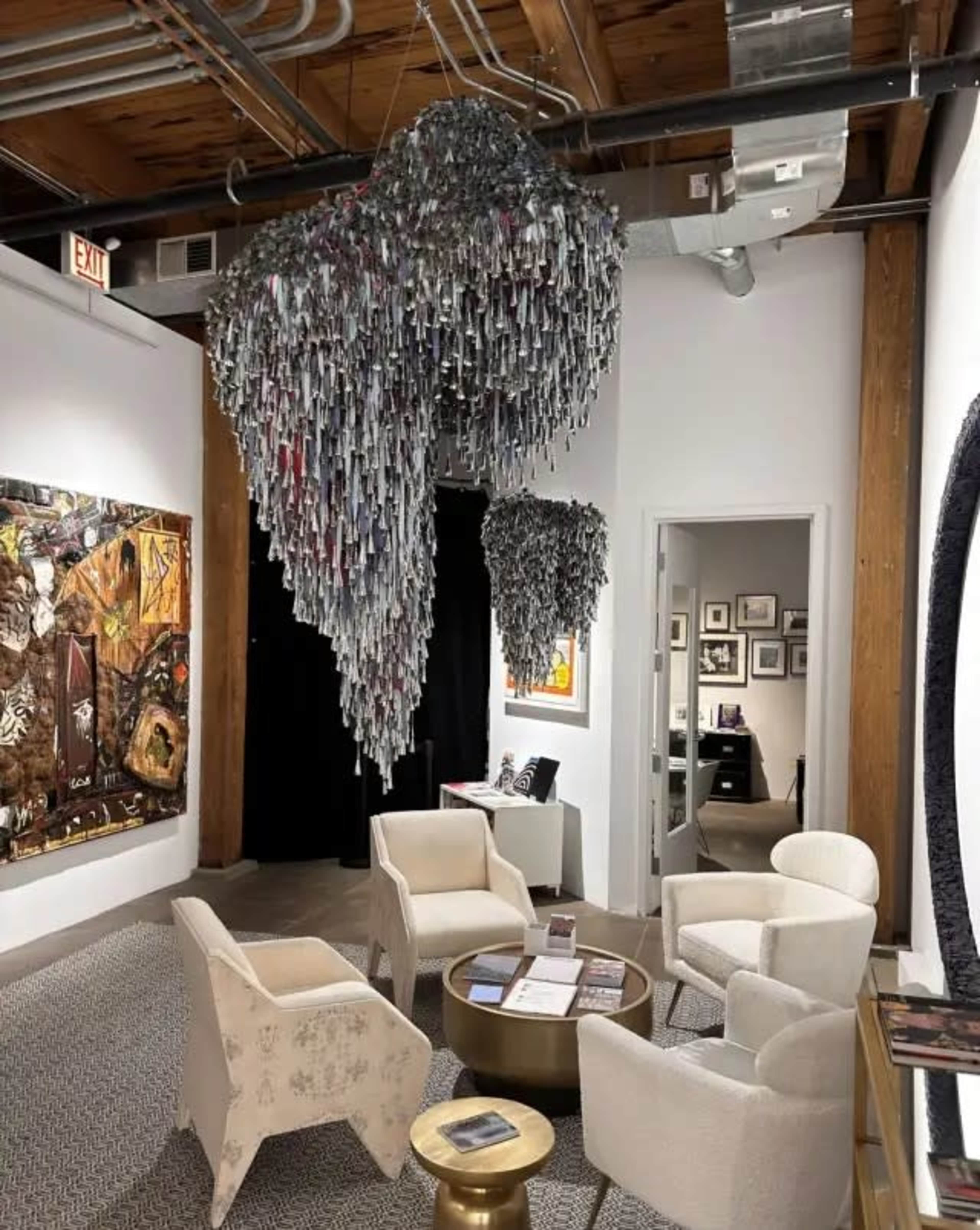 The image shows an art gallery with several chairs arranged around a circular table, featuring a large hanging sculpture made of elongated metallic materials overhead.