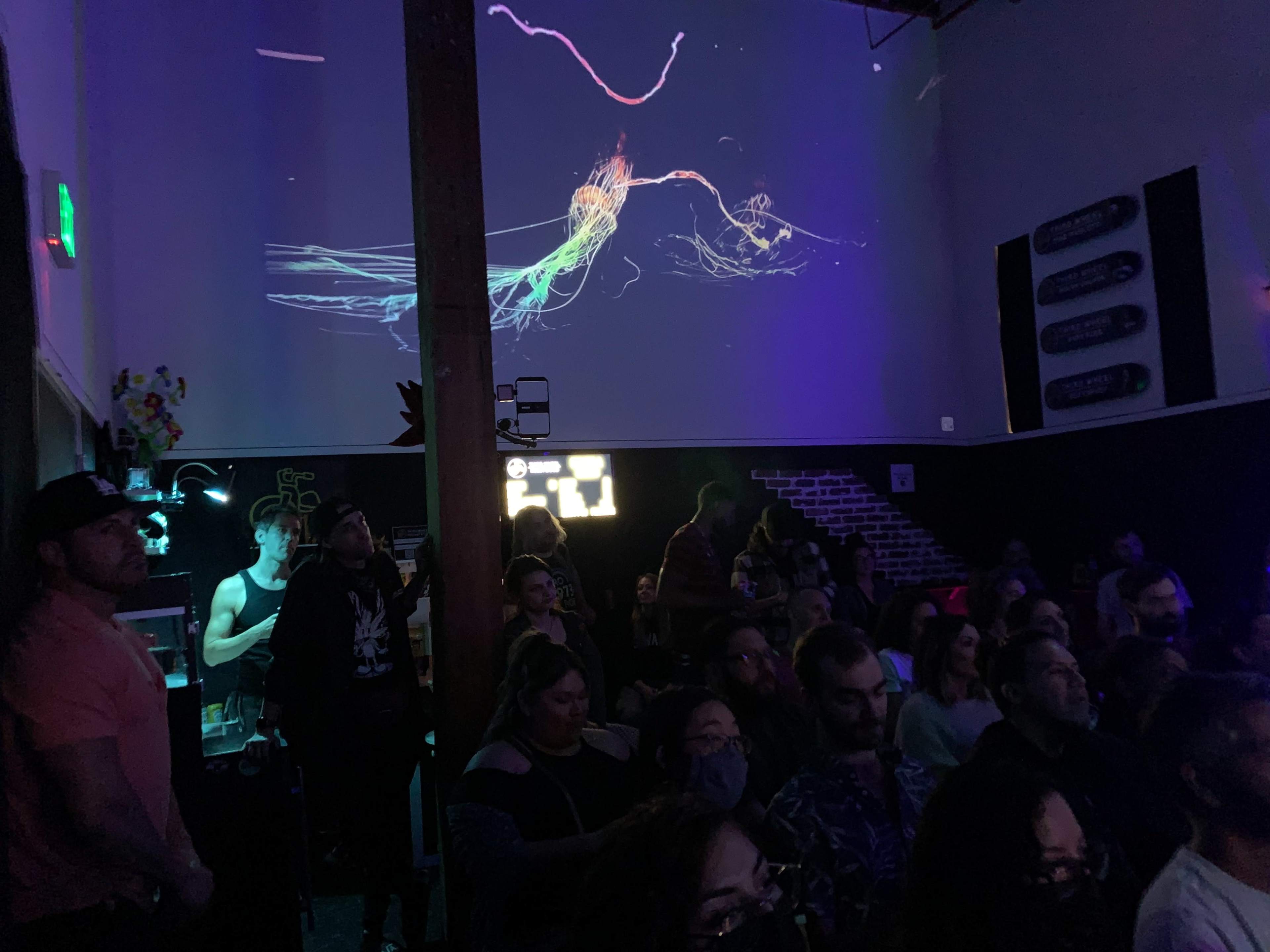 A crowd of people watches a colorful visual display projected on a wall in a dimly lit venue.
