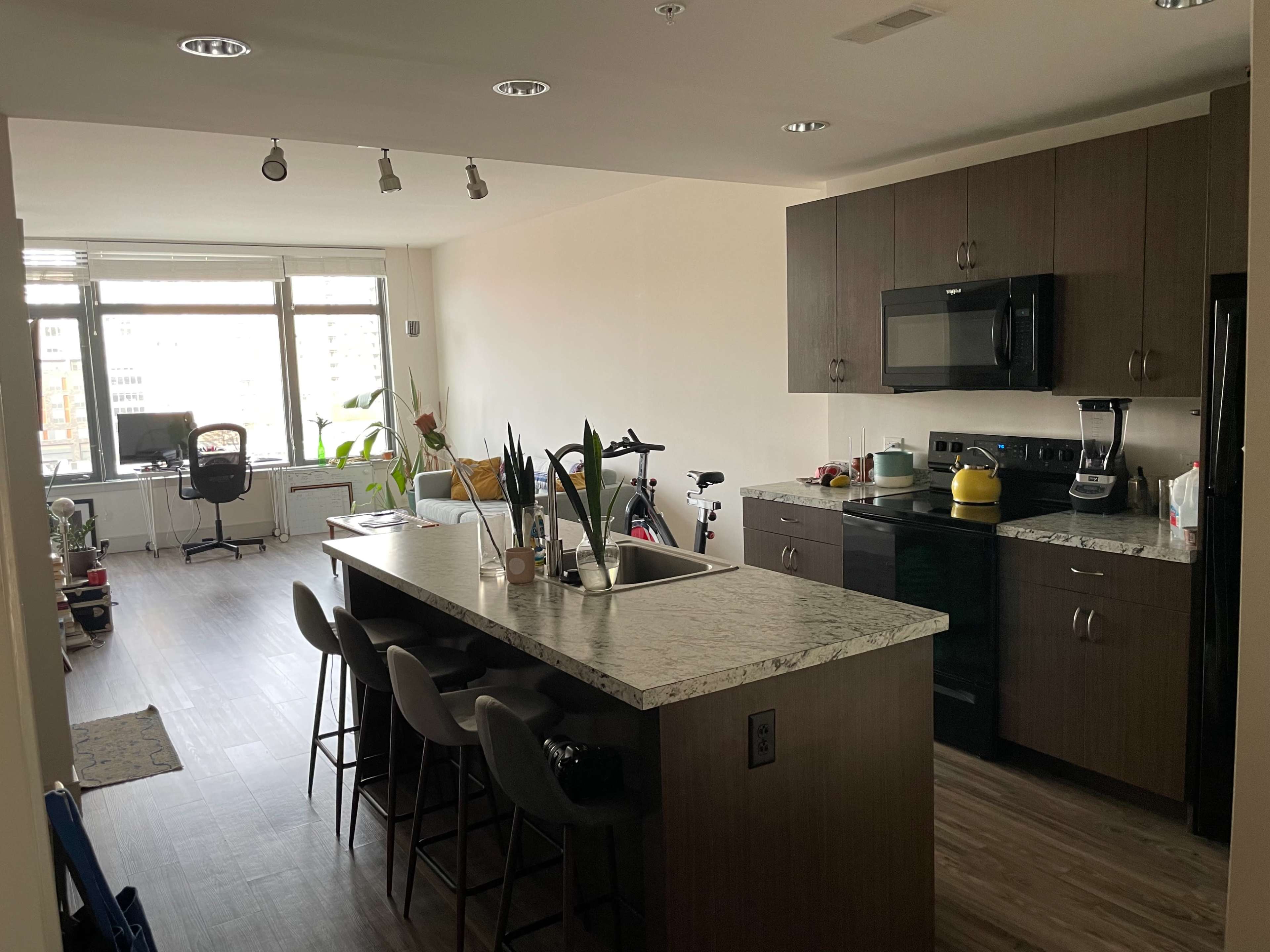 A modern kitchen with an island, four bar stools, and an adjacent living area featuring a desk and plants by the window.