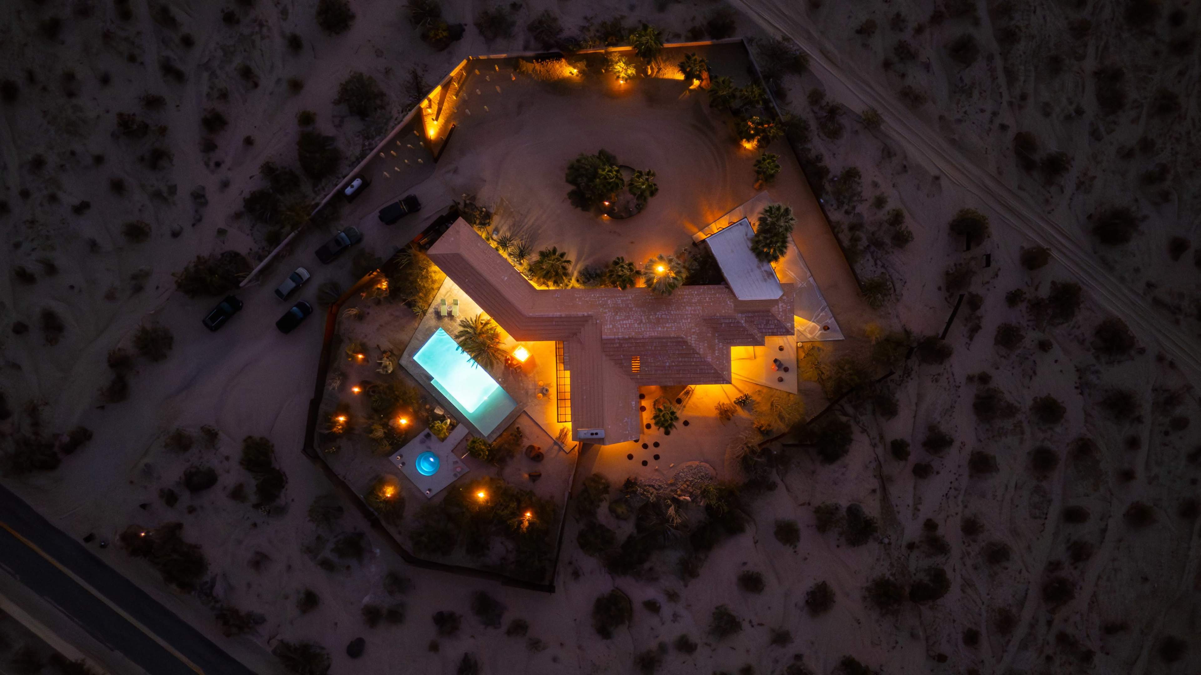 An aerial view of a modern desert house featuring a swimming pool, hot tub, and landscaping illuminated at night.