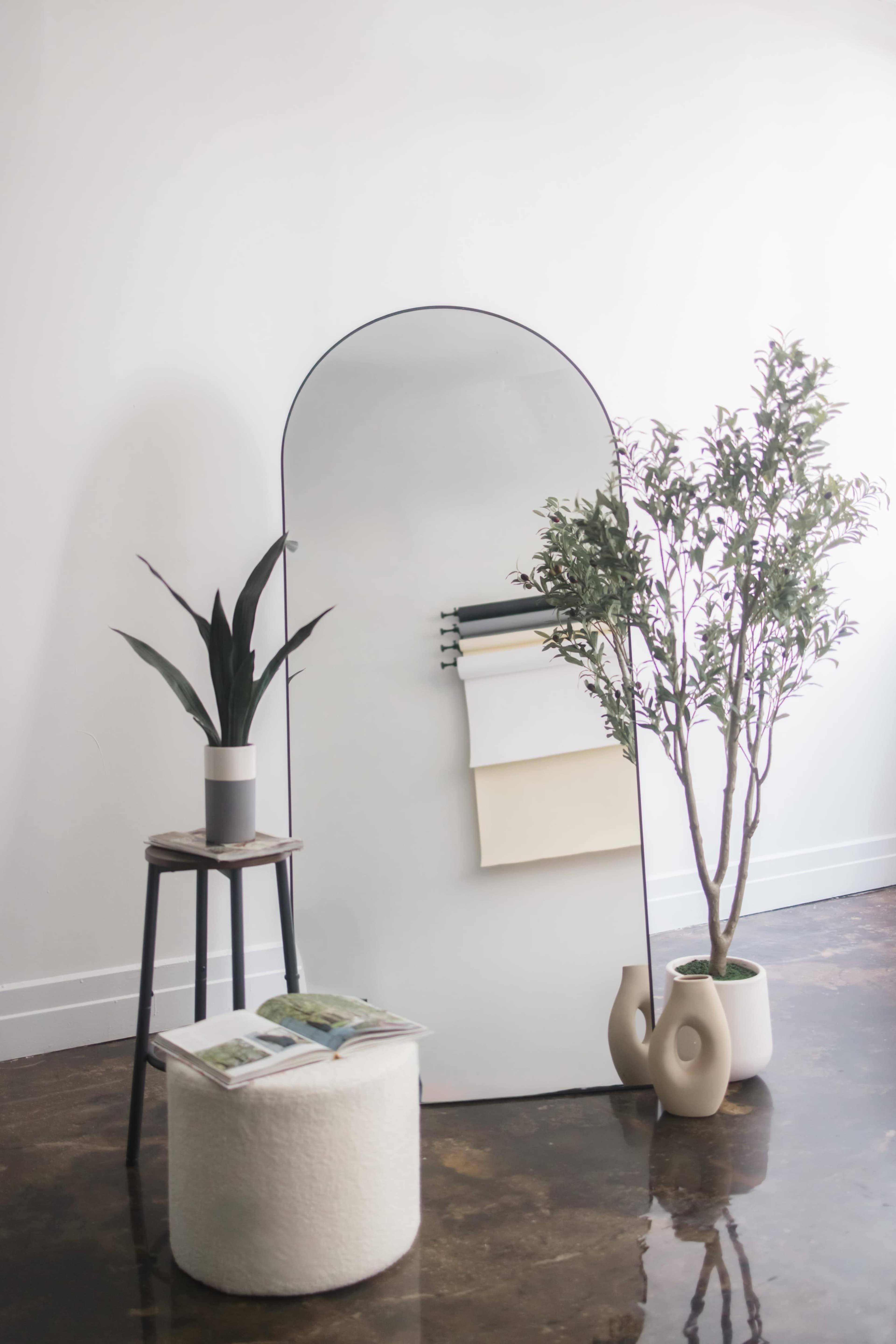 A tall mirror with an arched top stands against a white wall, surrounded by a plant on a stool, a potted tree, and a small, round pouf.