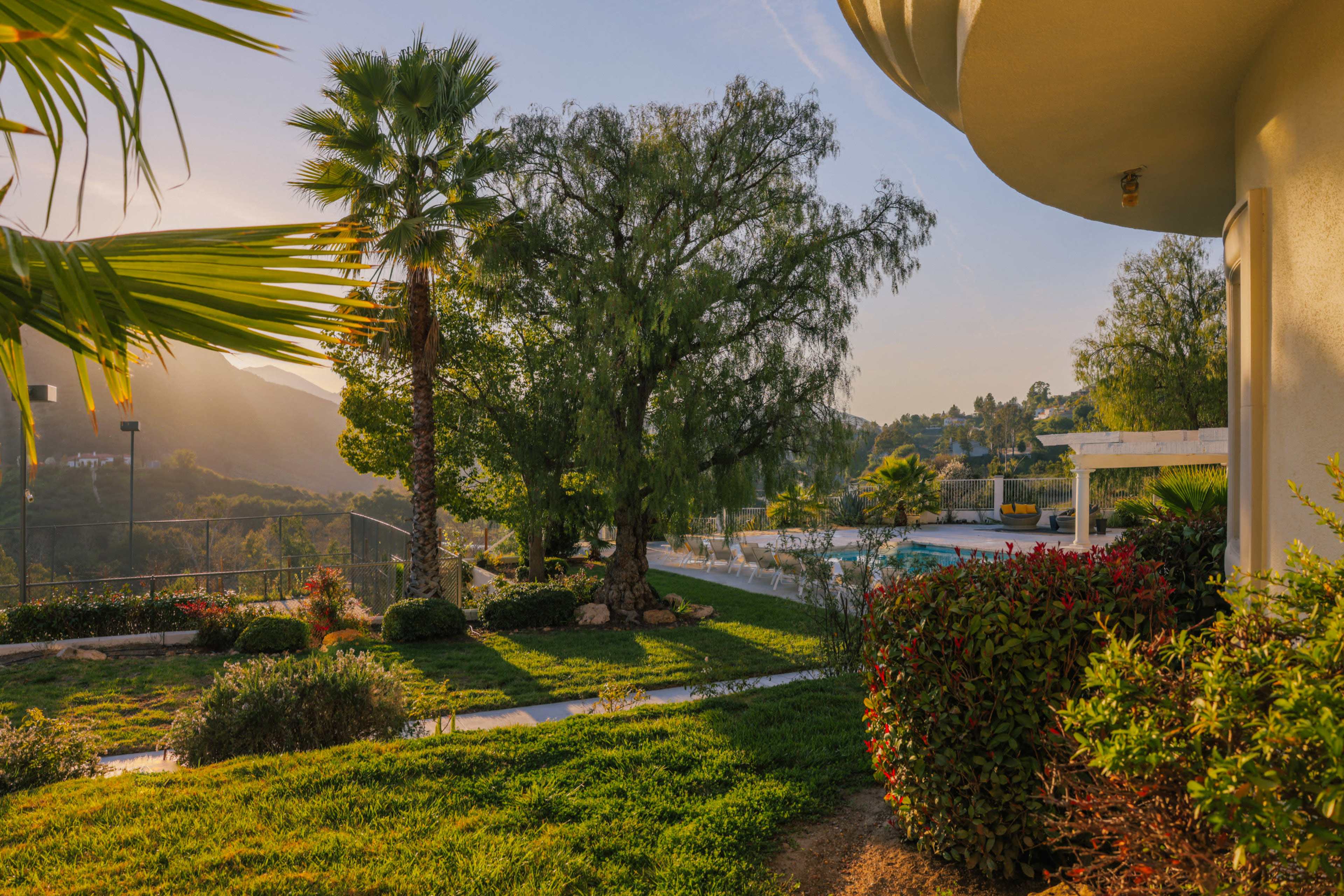 The scene features a landscaped garden with palm trees and a swimming pool surrounded by hills and greenery under a clear sky.