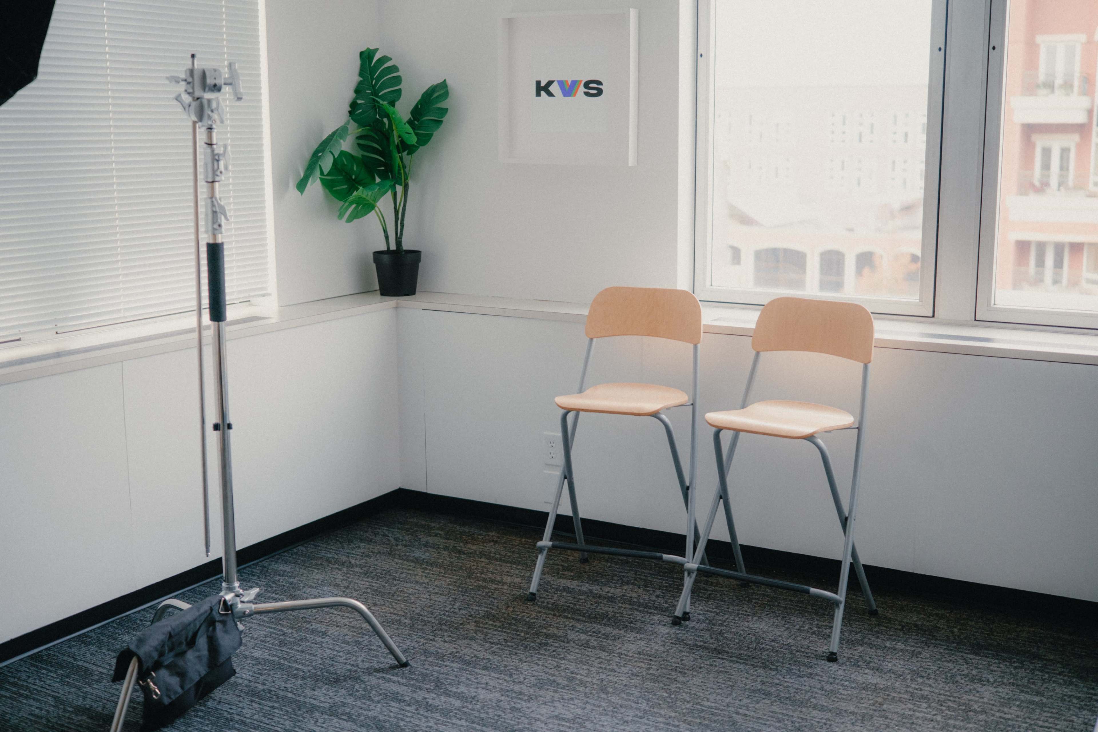 The image shows a minimalistic indoor setting with two wooden chairs, a potted plant, and a wall-mounted artwork featuring the letters "KVS" near a large window.