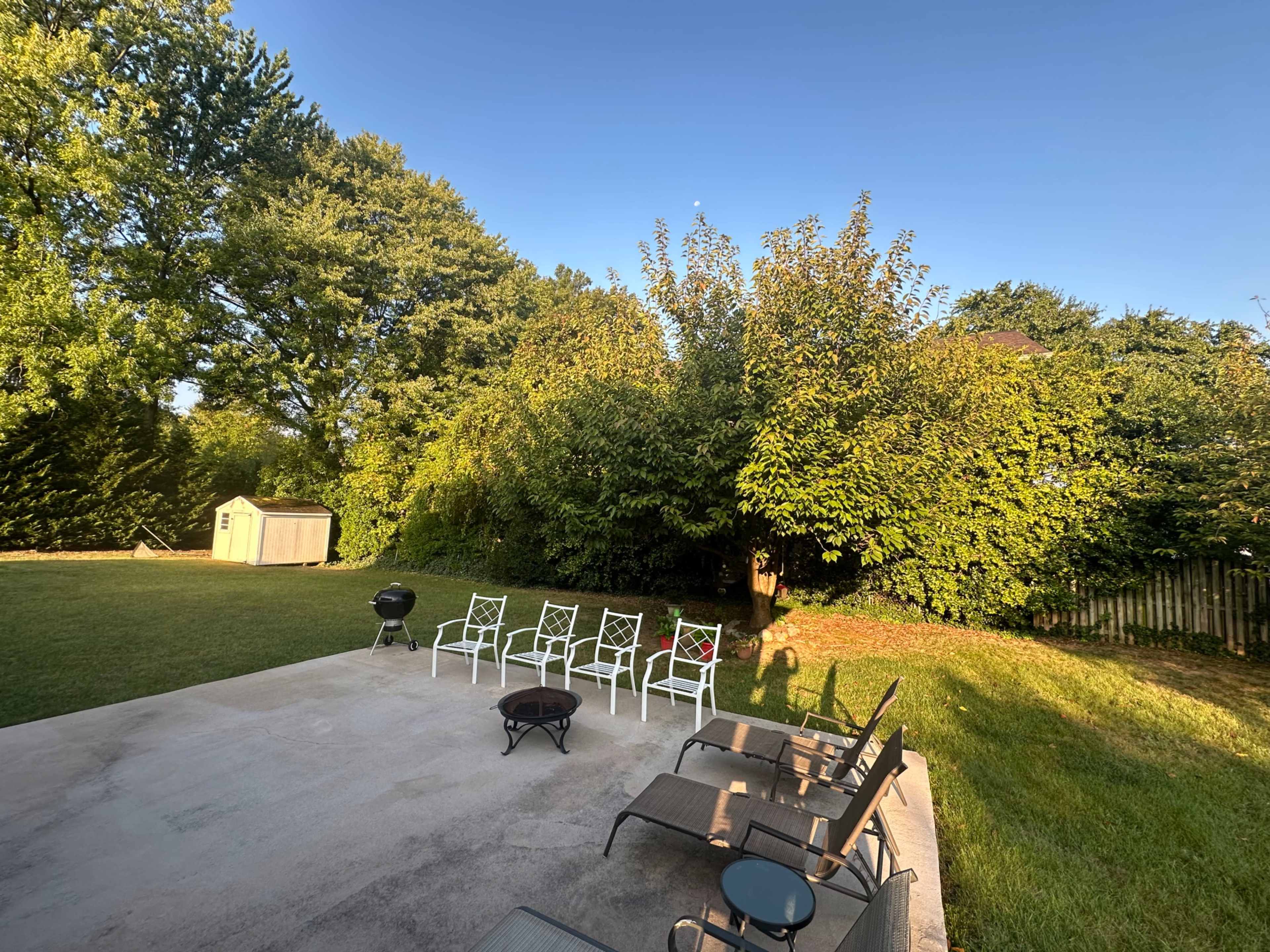 A patio area with white chairs and a small table overlooks a green lawn and dense trees, with a storage shed visible in the background.