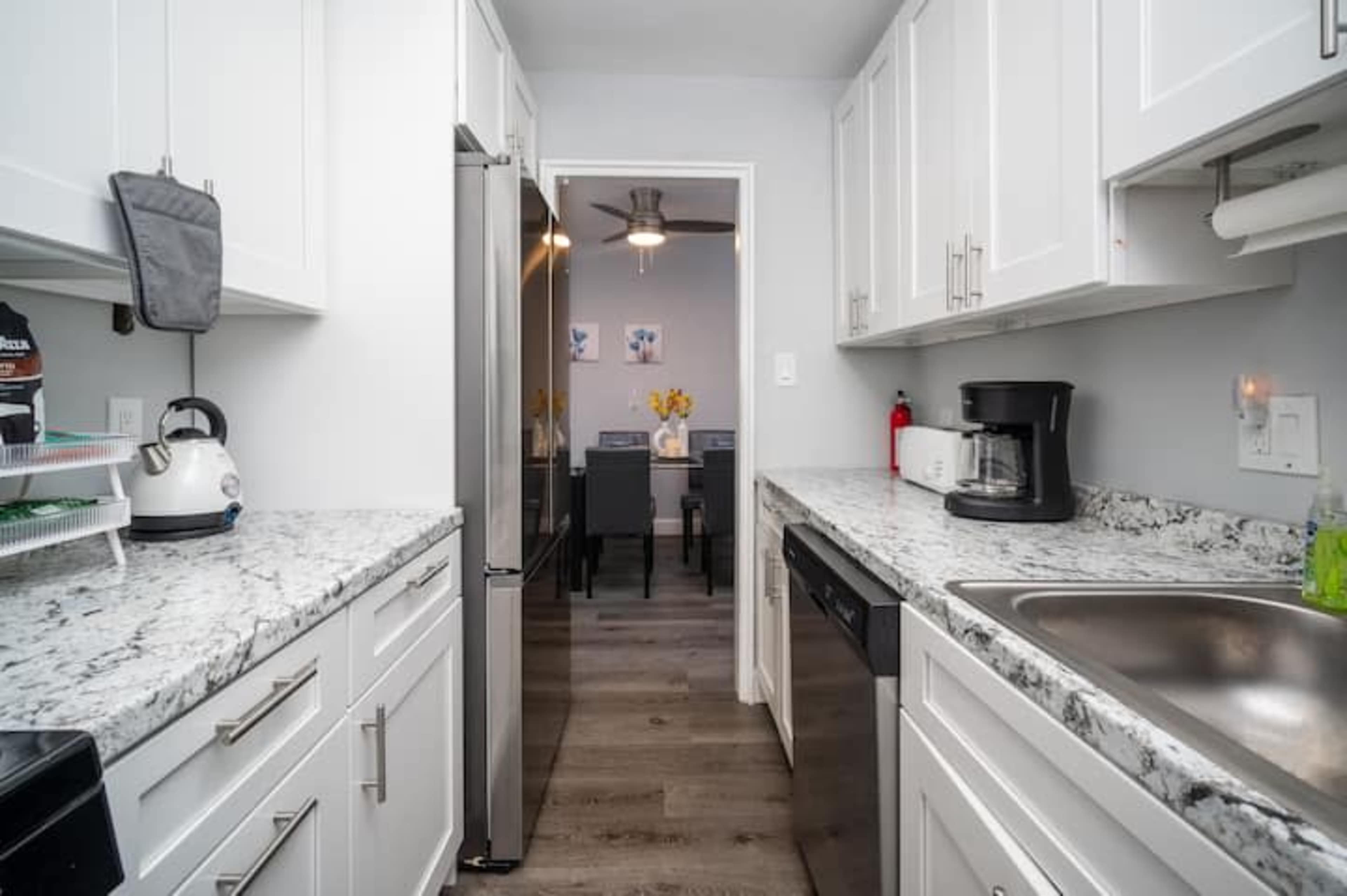 A modern kitchen with white cabinets, a granite countertop, and stainless steel appliances, leading to a dining area in the background.