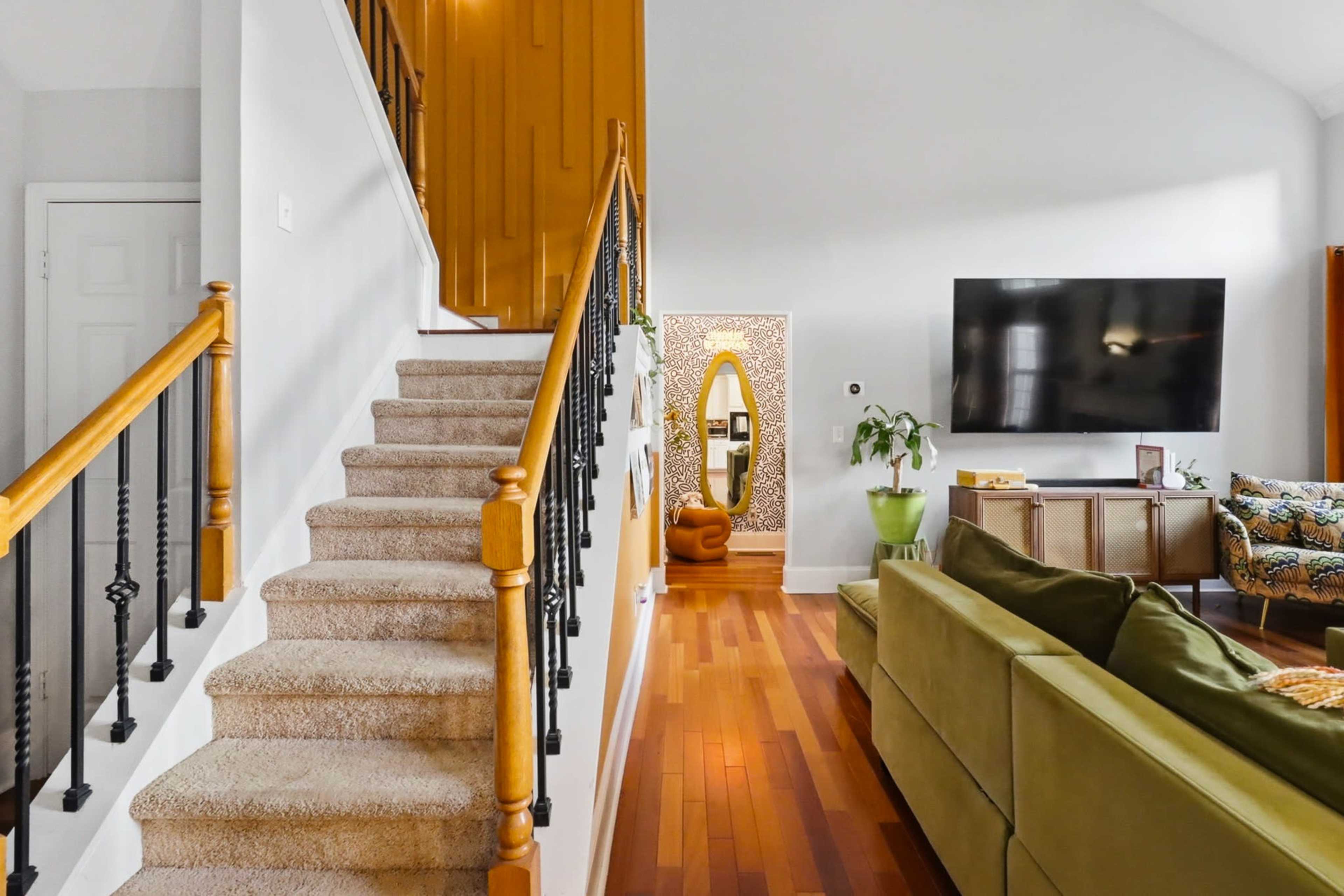 The image shows a well-lit interior space featuring a staircase on the left, a living area with a green couch, and a television mounted on the wall.