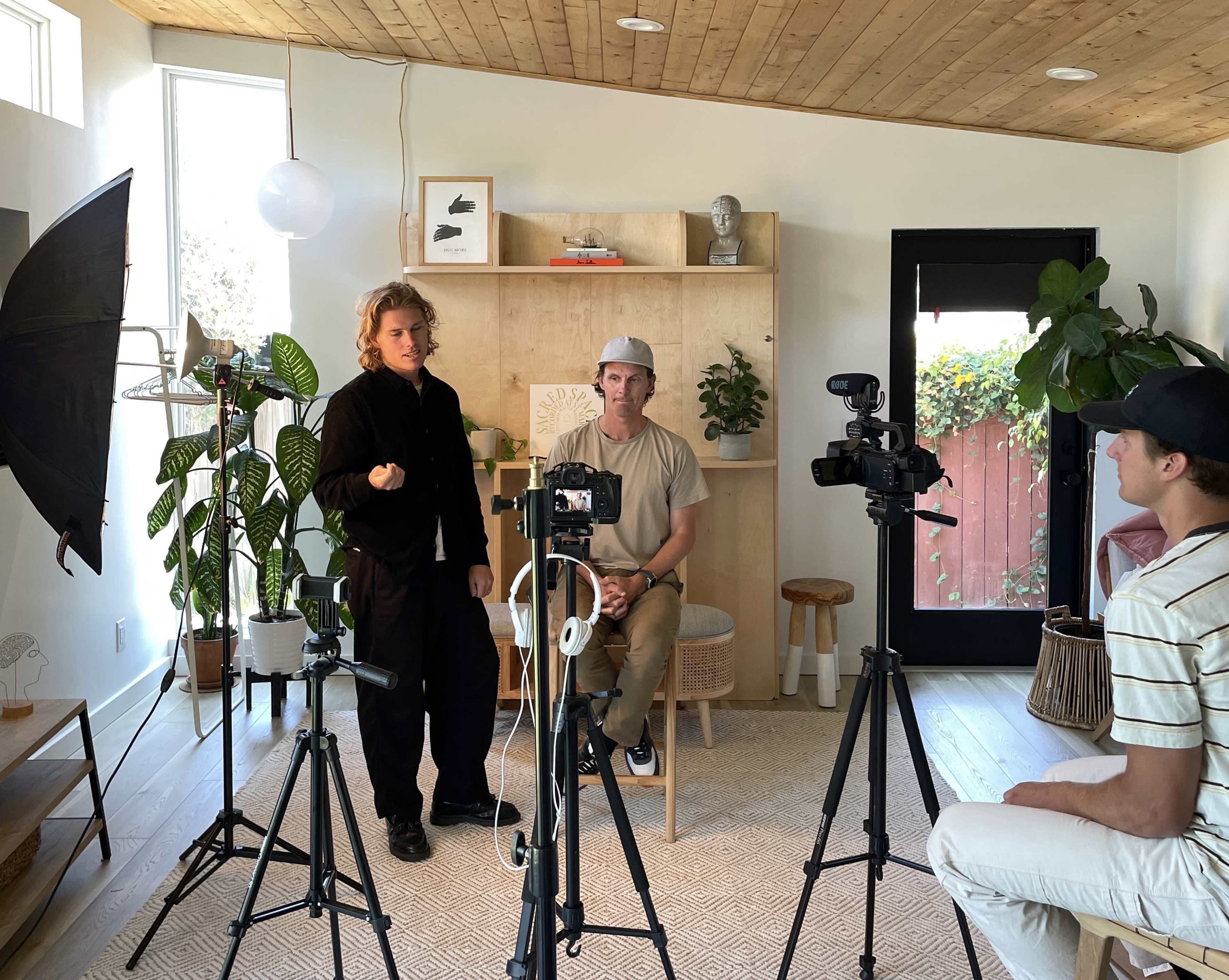 Two people are positioned in front of cameras in a well-lit room with a wooden ceiling, while a third person sits in a chair, and plants decorate the space.