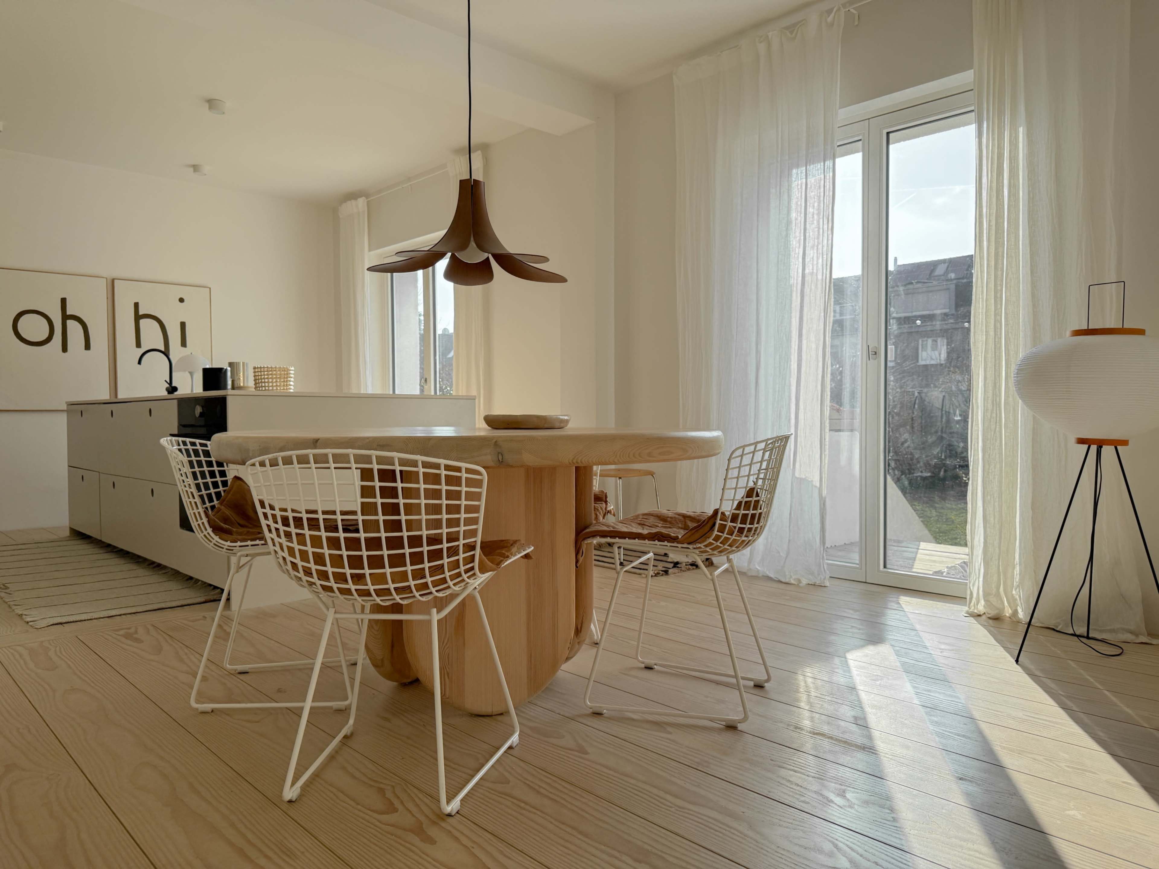 A round wooden dining table surrounded by wire chairs is positioned near a large window in a bright, minimalist room.
