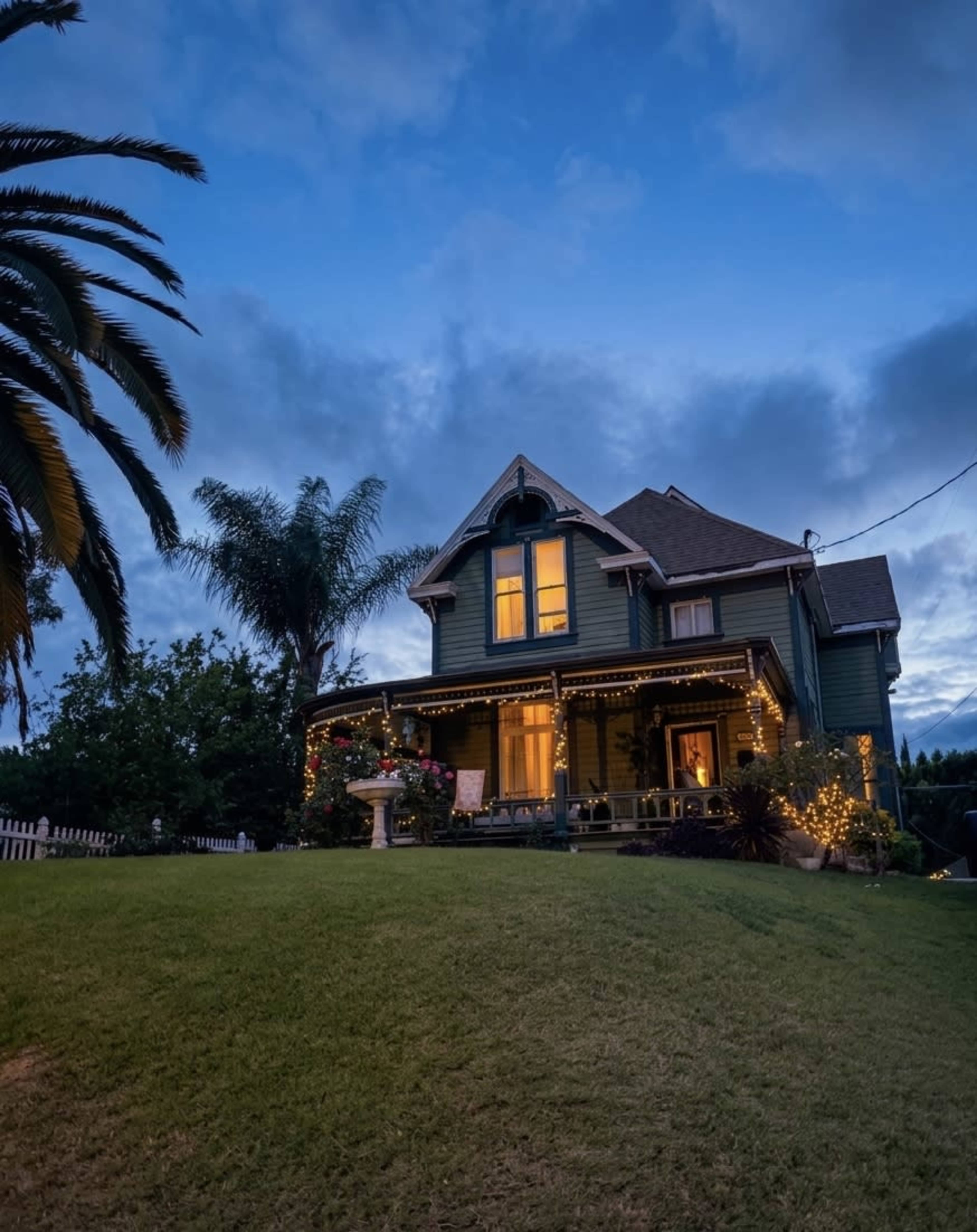 A two-story house with decorative lights is set on a grassy lawn under a twilight sky, surrounded by palm trees and shrubbery.