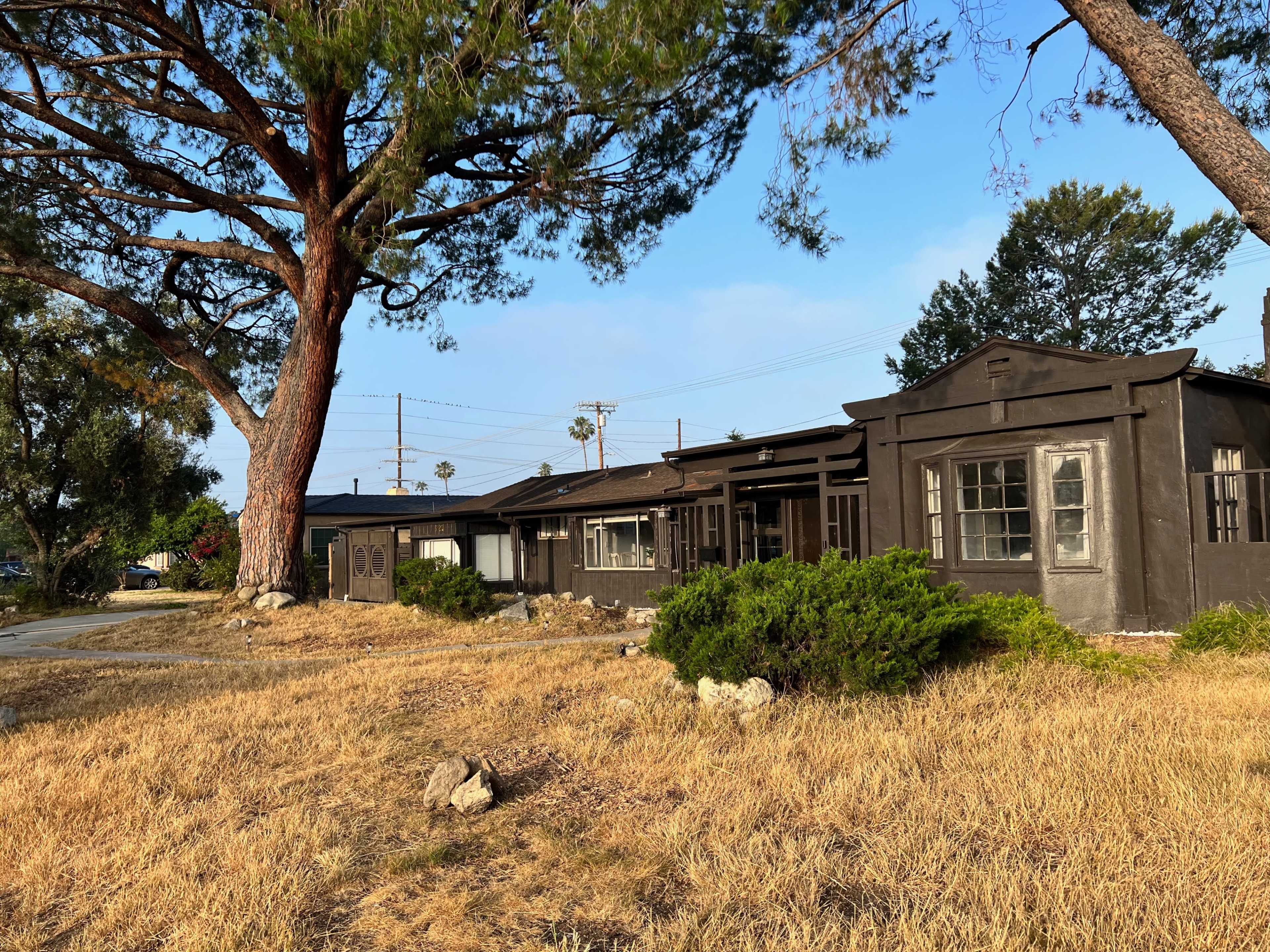 A weathered, dark-colored house with a porch is surrounded by dry grass and trees in a suburban area.