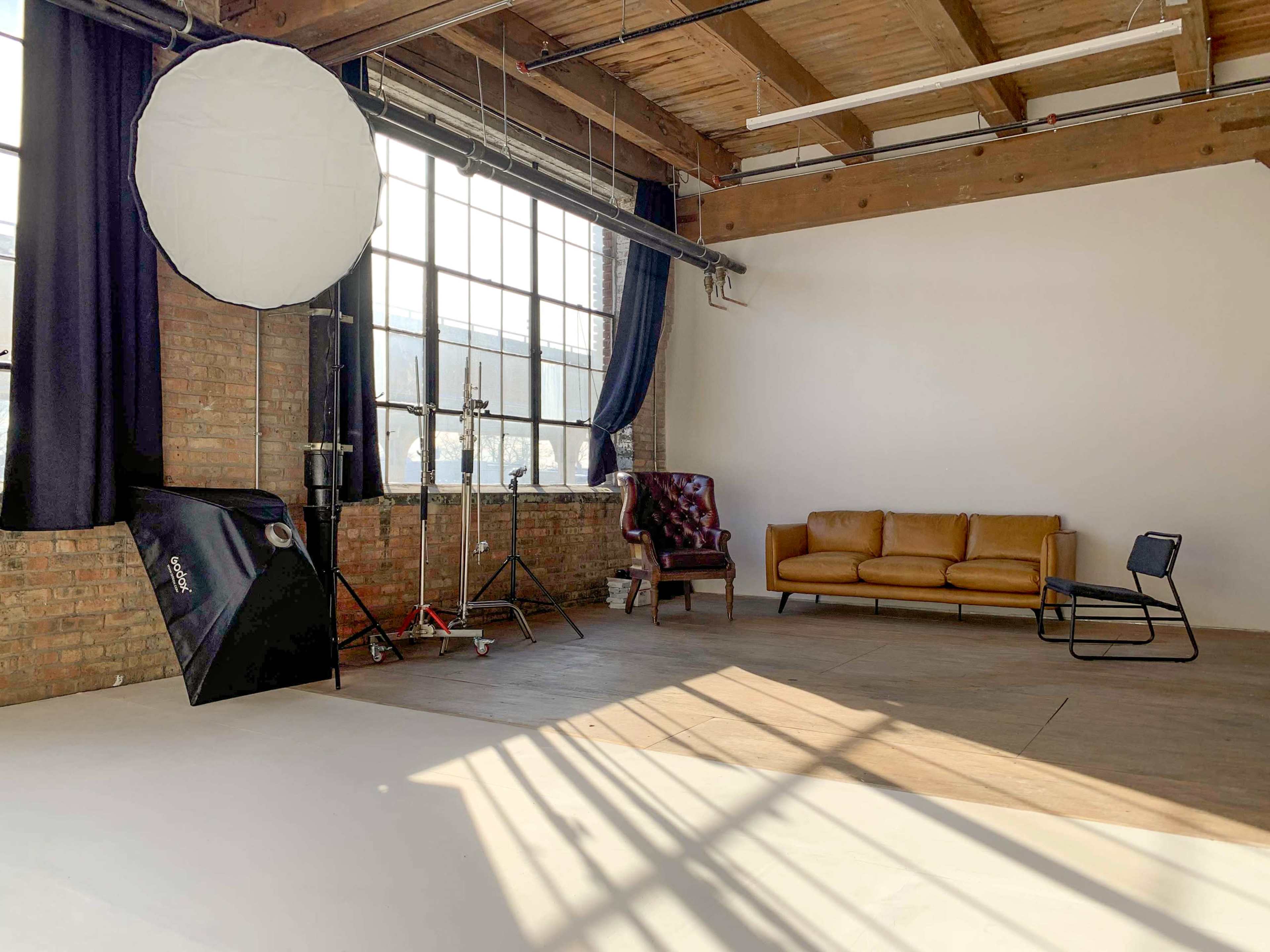 A spacious studio with large windows, featuring a brown leather couch, a single chair, and a light reflector on a stand.
