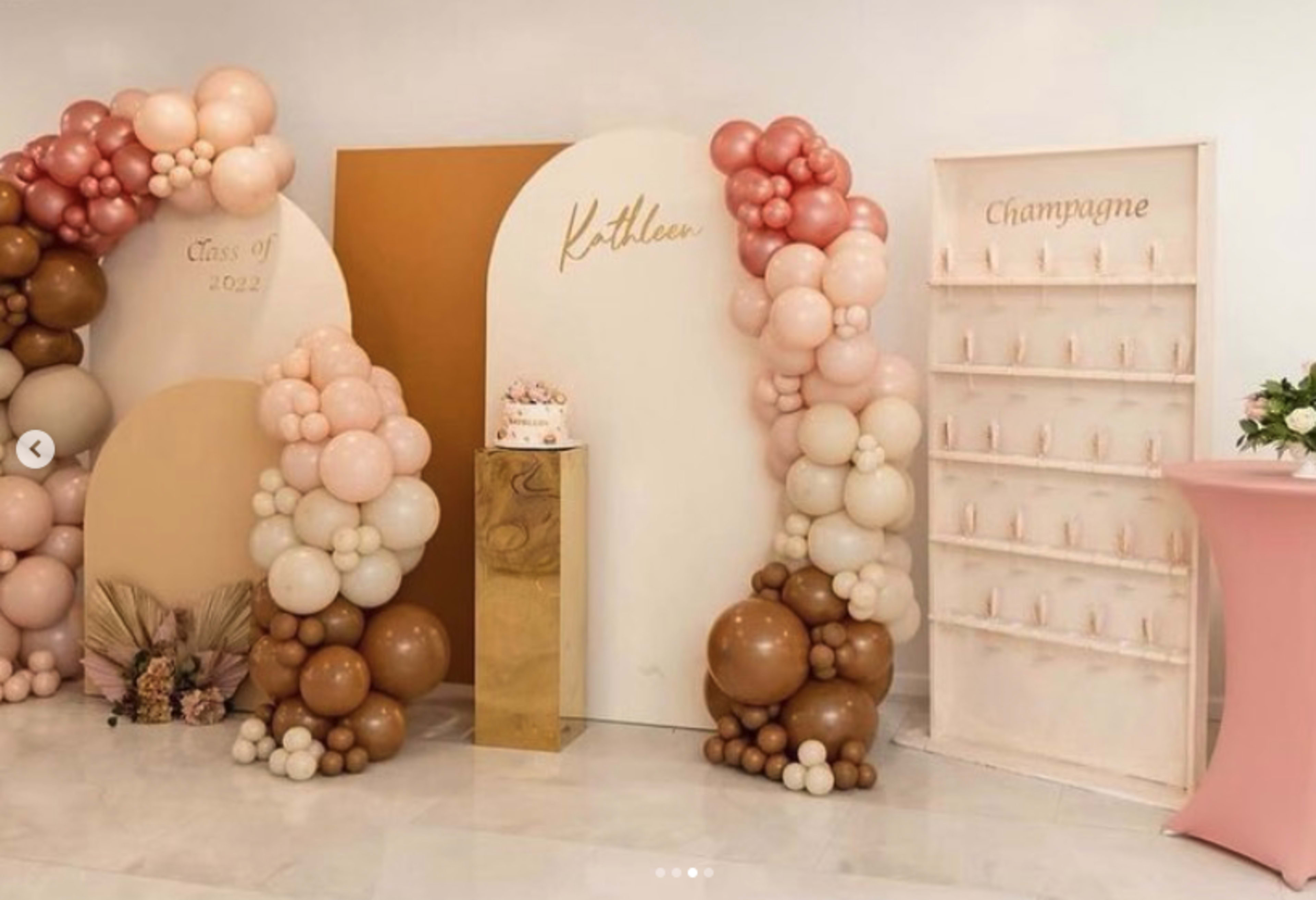 The scene features a festive decor setup with balloon arches, a gold pedestal displaying a cake, and a champagne wall, all arranged for a graduation celebration.