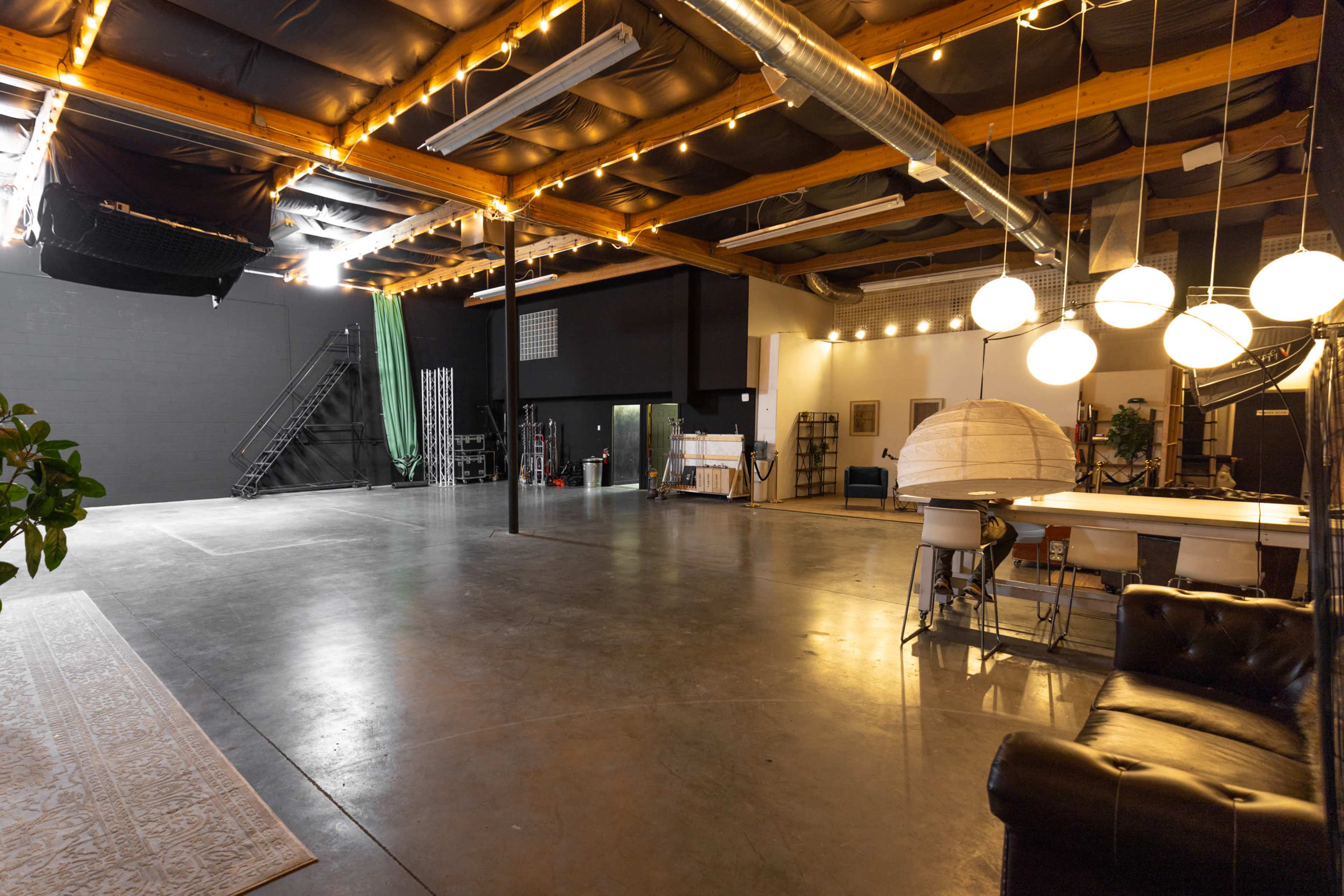 The image shows a spacious studio with high ceilings, concrete flooring, and various equipment, including a couch, lighting fixtures, and a staircase in the background.
