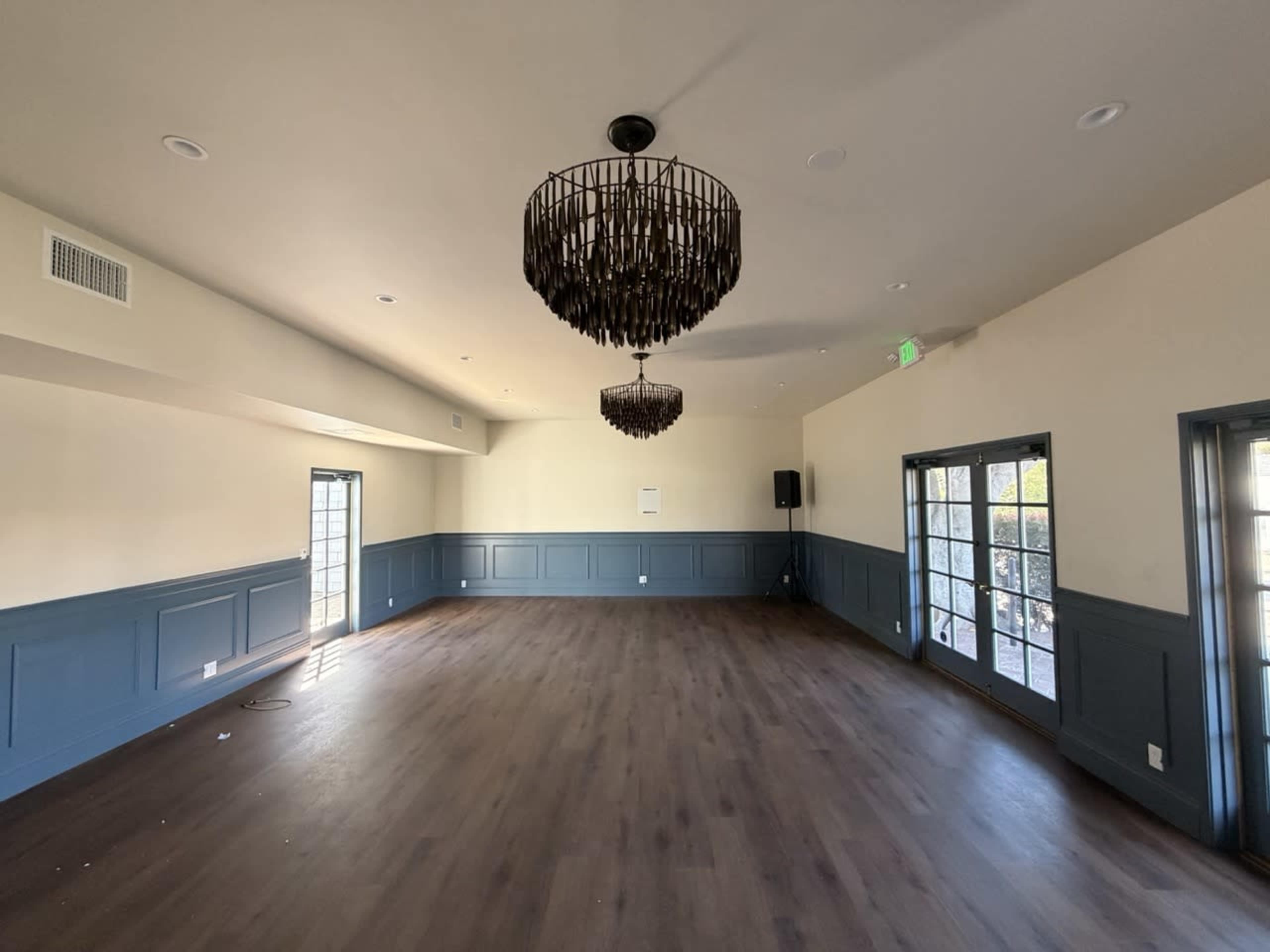 The image shows an empty room with wooden flooring, two large chandeliers, and blue wainscoting on the walls, featuring multiple windows and doors.