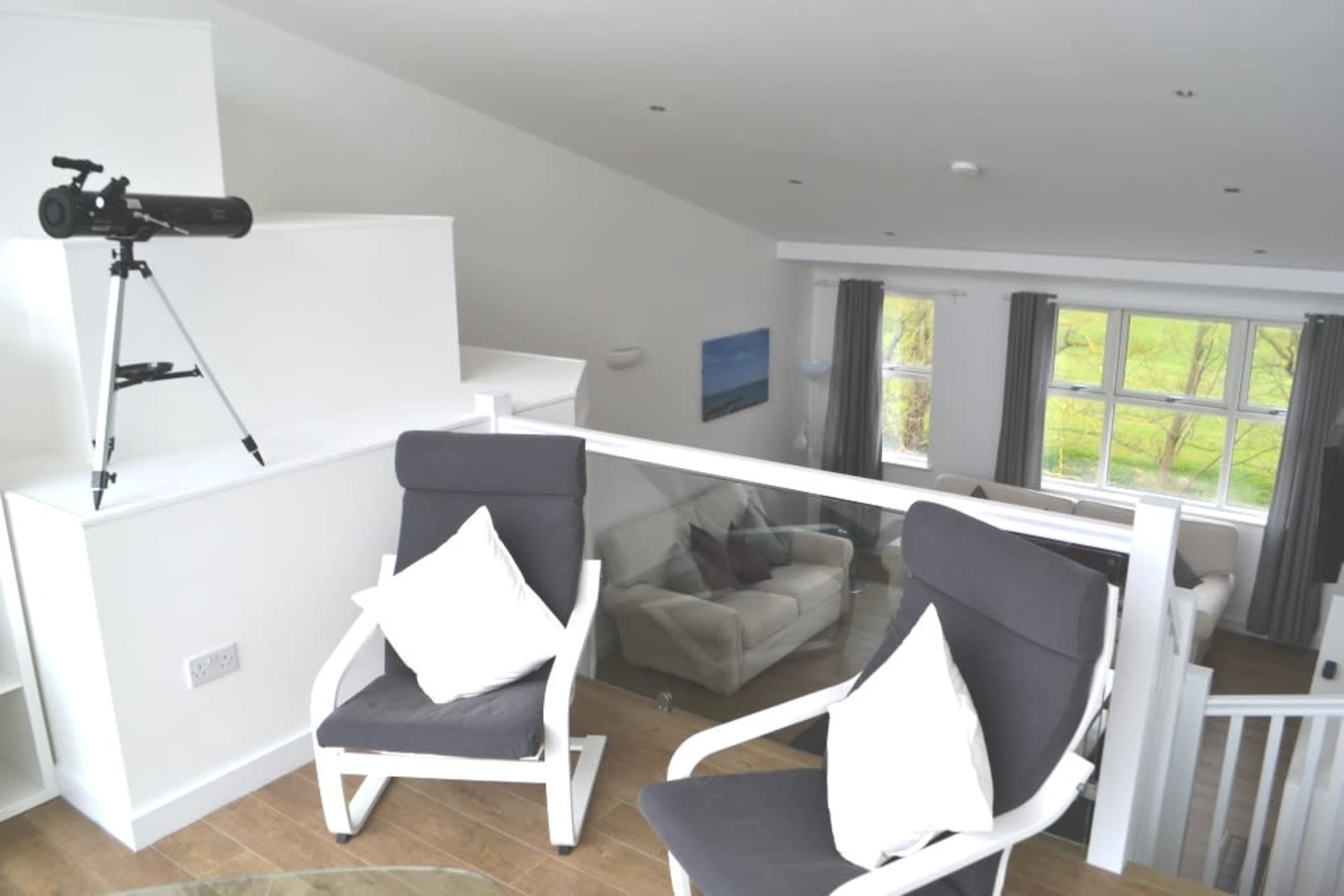 The image shows a spacious, modern interior featuring two white chairs with cushions, a telescope positioned on a stand, and large windows letting in natural light.
