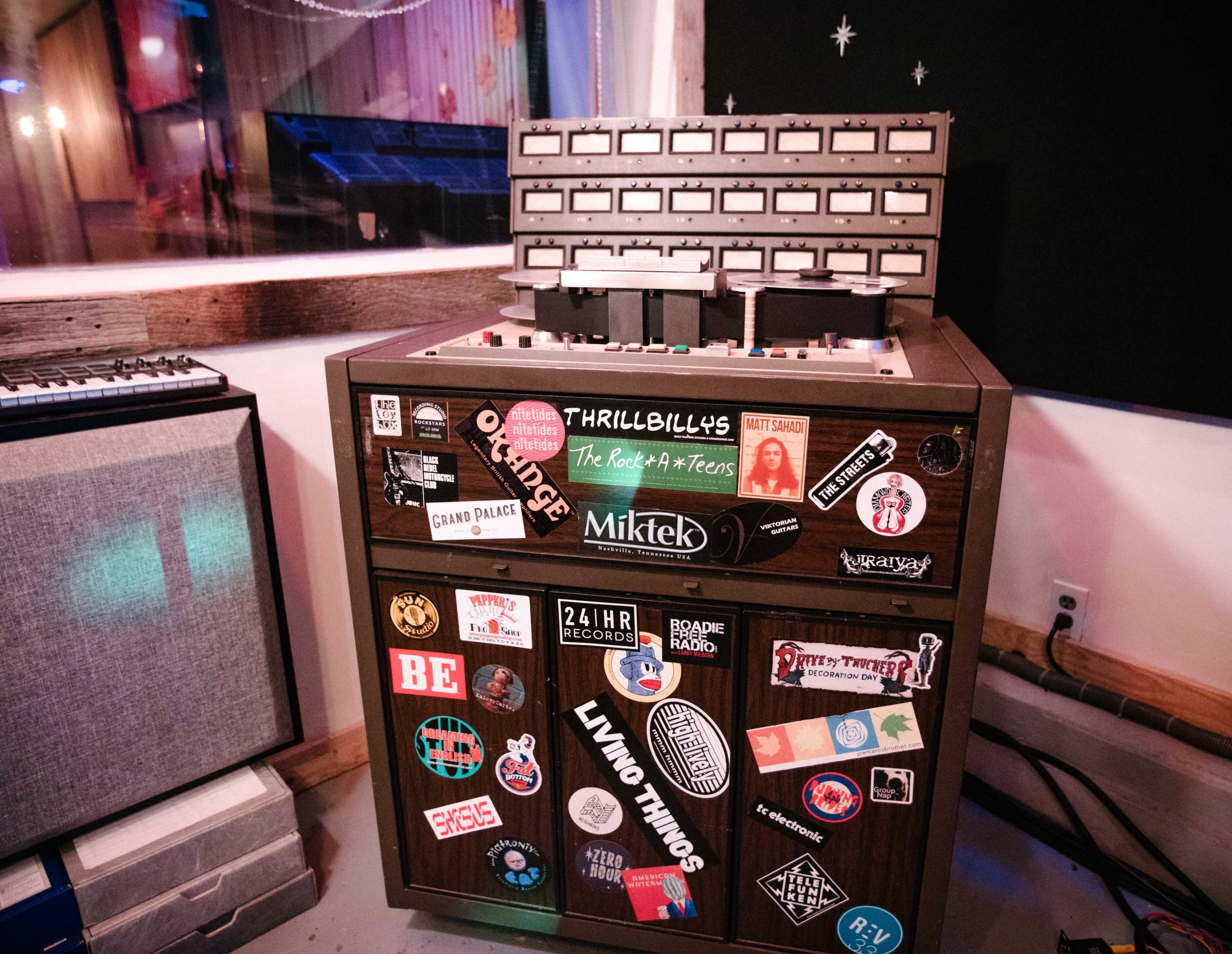 A wooden audio mixing console adorned with various colorful stickers, positioned in a room with musical equipment.