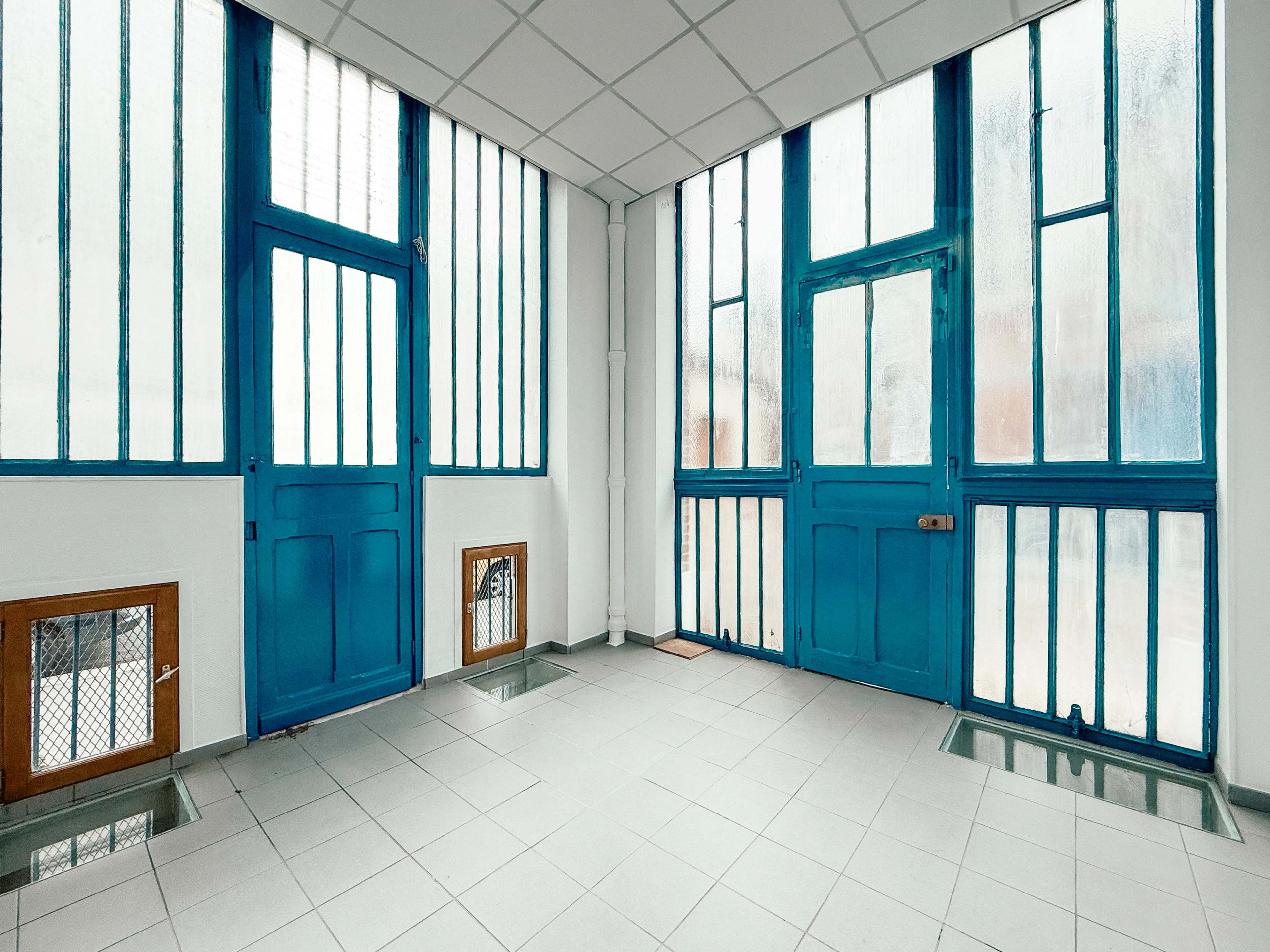 The image shows a bright, airy interior space with large windows and two blue doors, highlighted by a tiled floor and minimal furnishings.