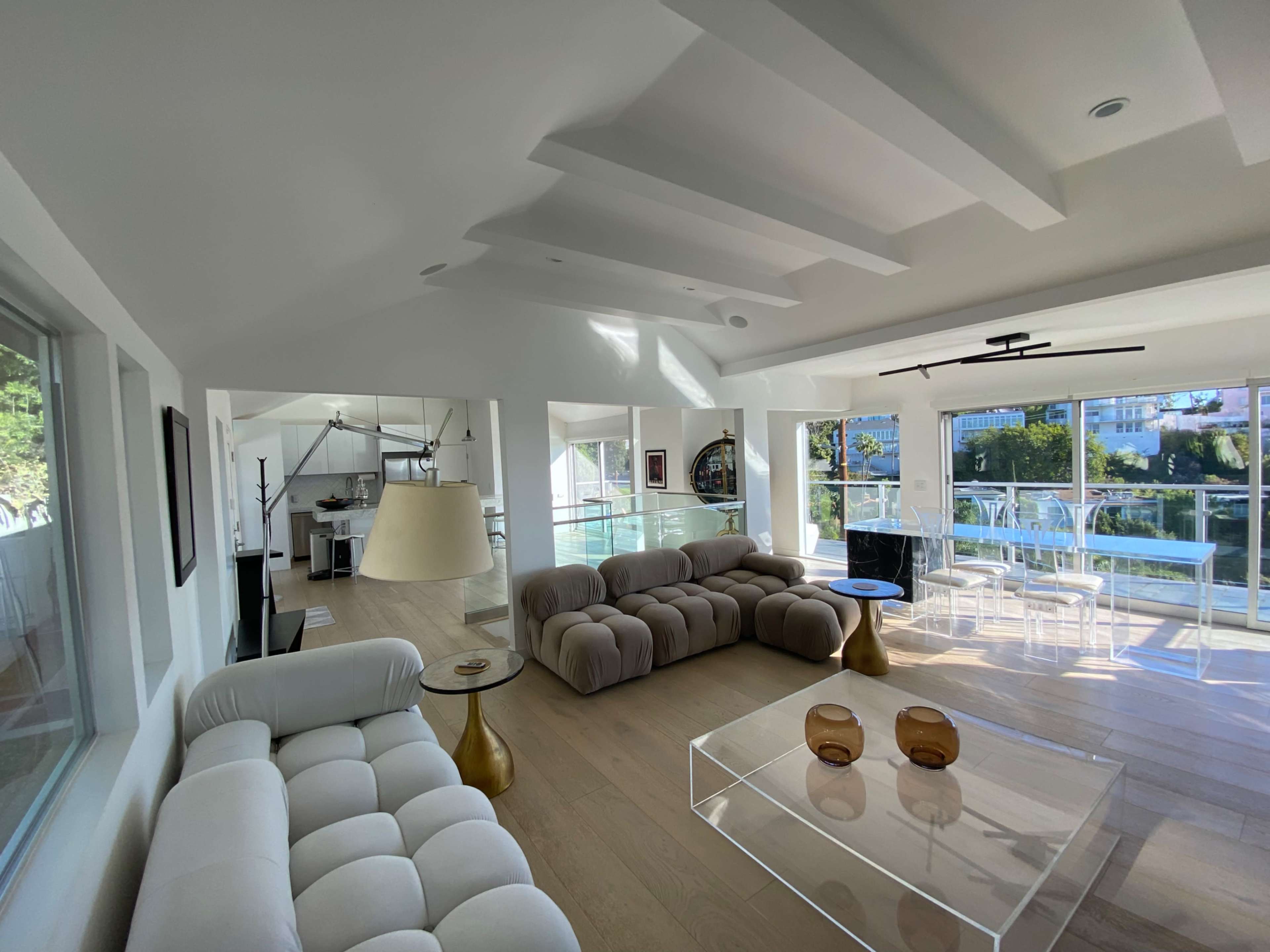 The image shows a modern open-concept living area featuring two sofas, a glass coffee table, and large windows that provide a view of the outdoor landscape.