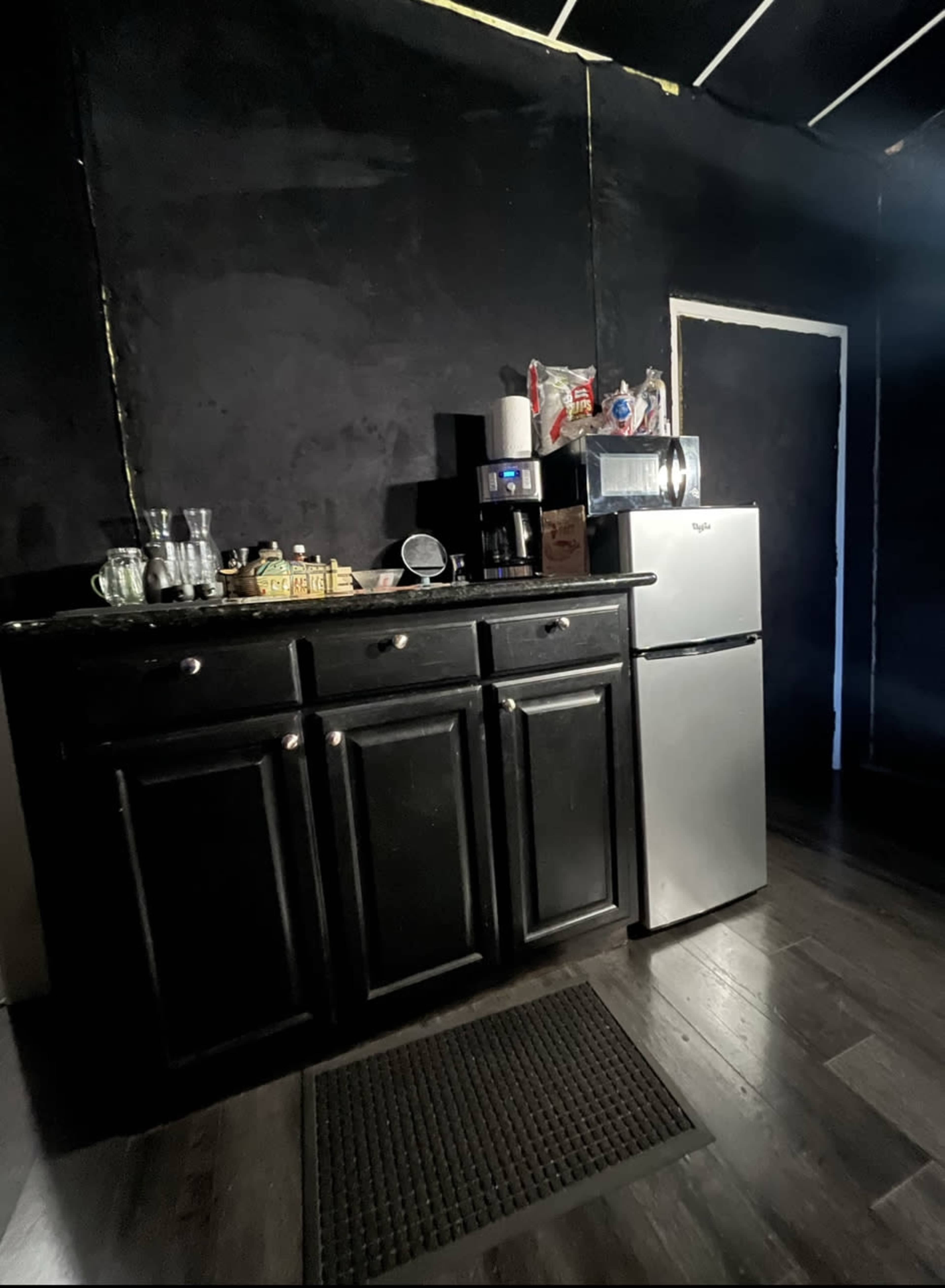 A small kitchen with black walls, a dark cabinet featuring a sink and several glassware items, and a silver refrigerator next to a white door.