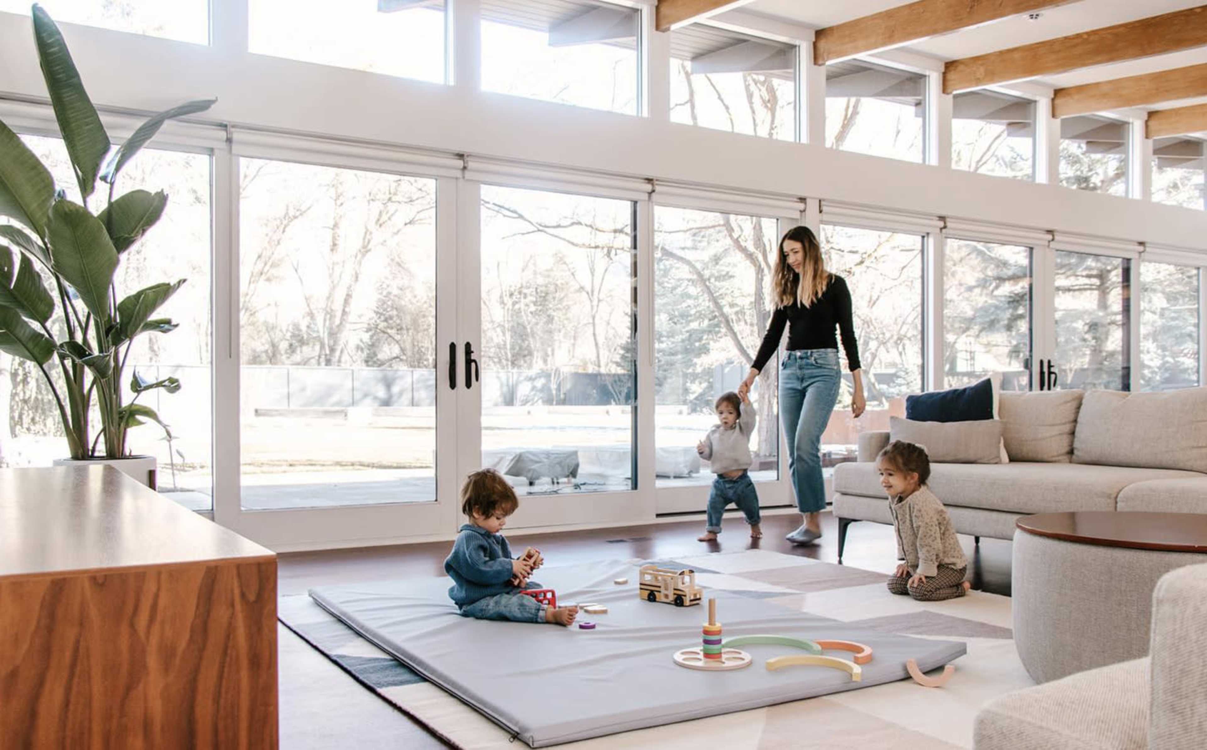 A woman stands beside a play area in a bright living room with large windows, while three children play on a mat.