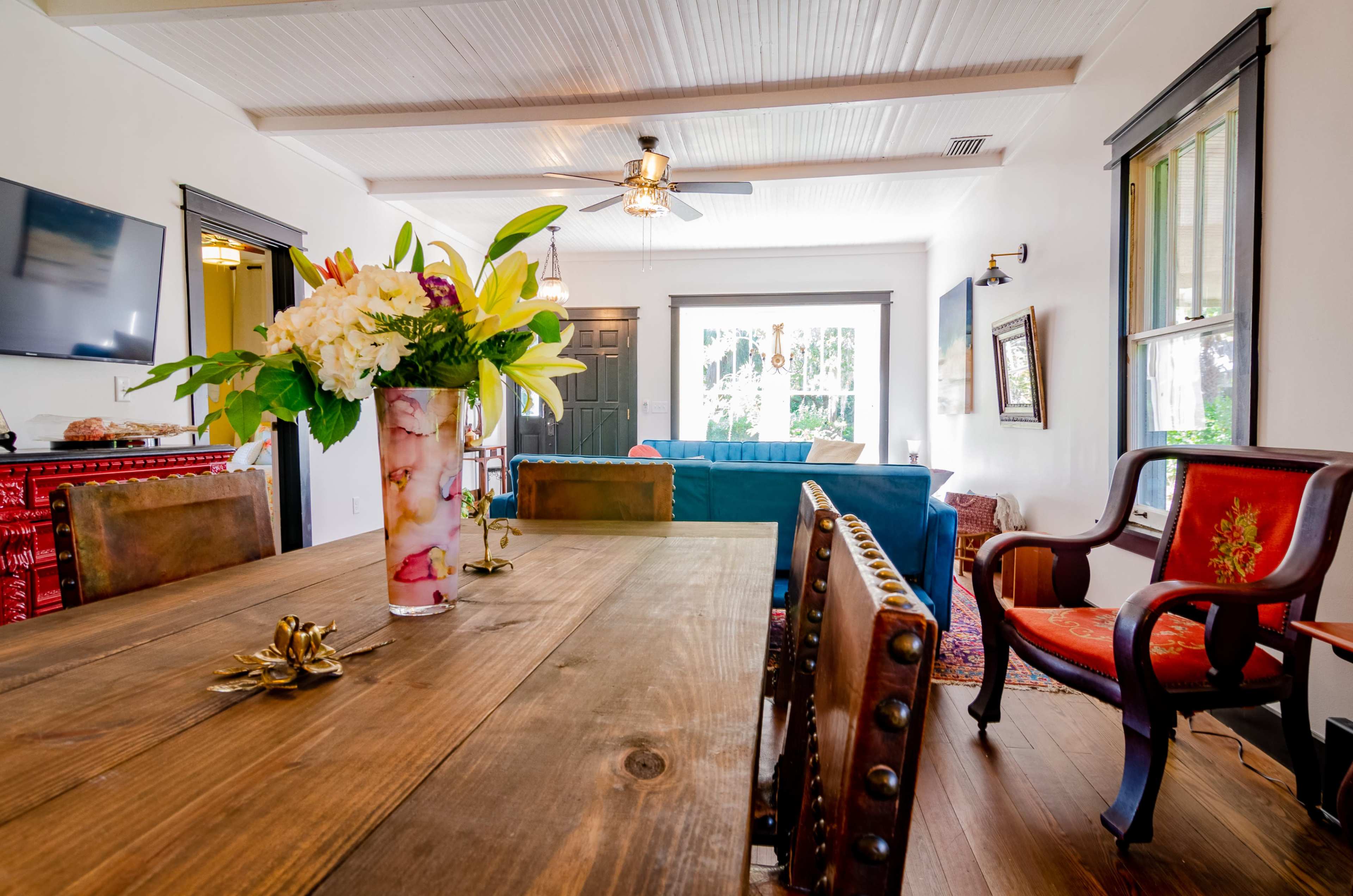 A dining area with a wooden table adorned with a floral arrangement, surrounded by chairs, and featuring a view of a living room with a blue couch and large windows.