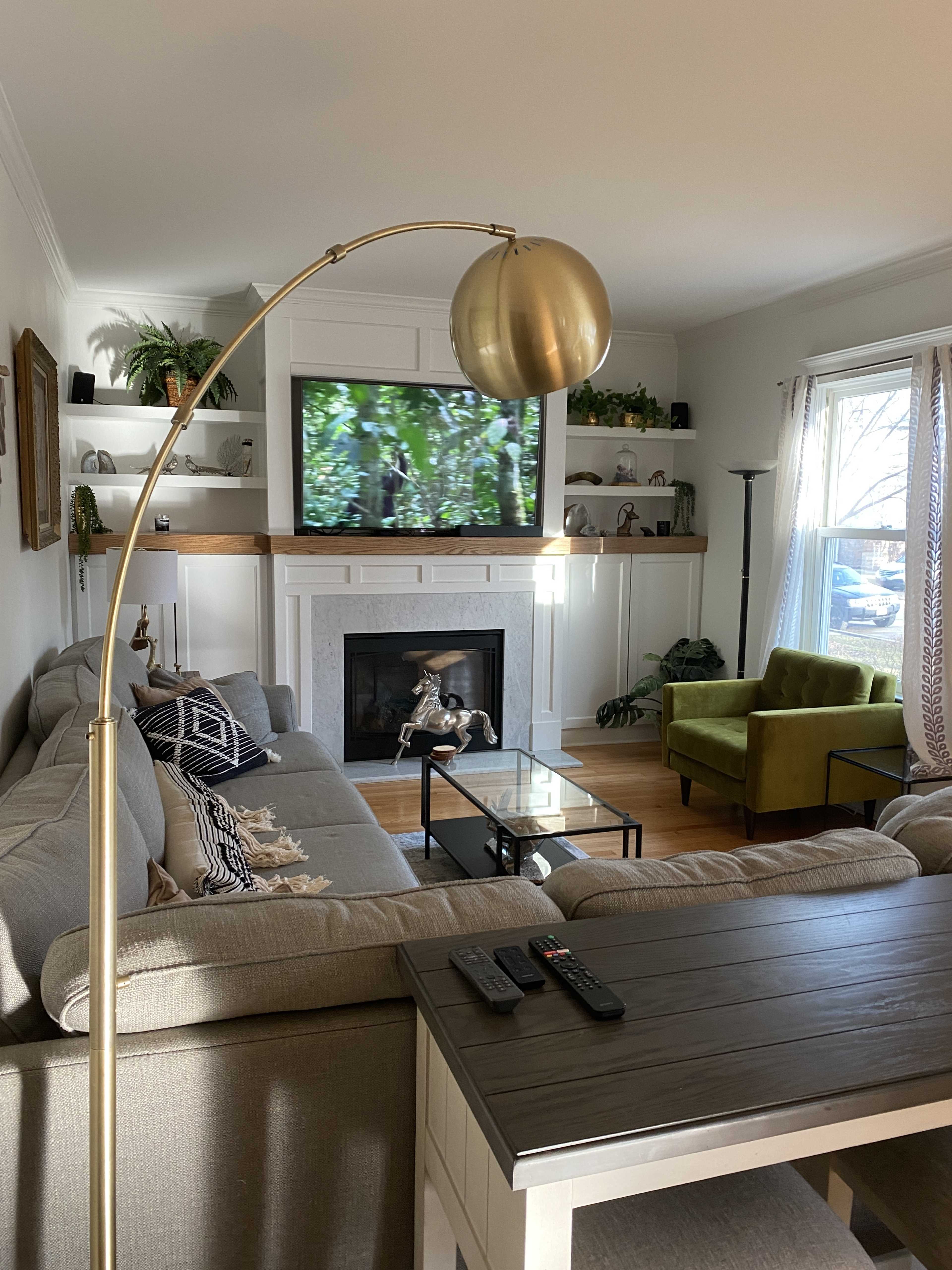 The living room features a sectional sofa, a coffee table, and a large television mounted above a fireplace, with greenery visible through the window.