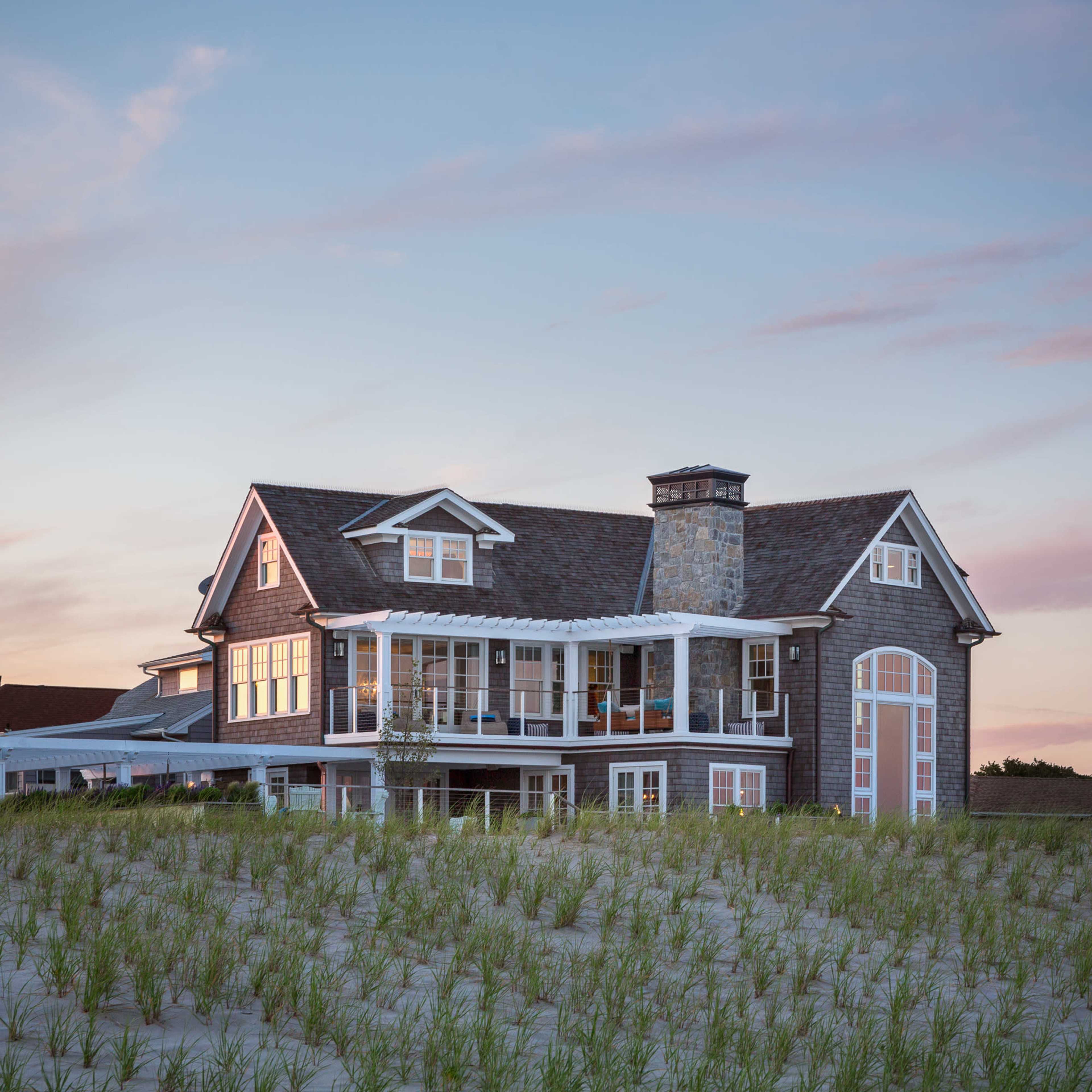 A large, coastal house with a gabled roof and multiple balconies is set against a backdrop of a pastel sky and beach grass.