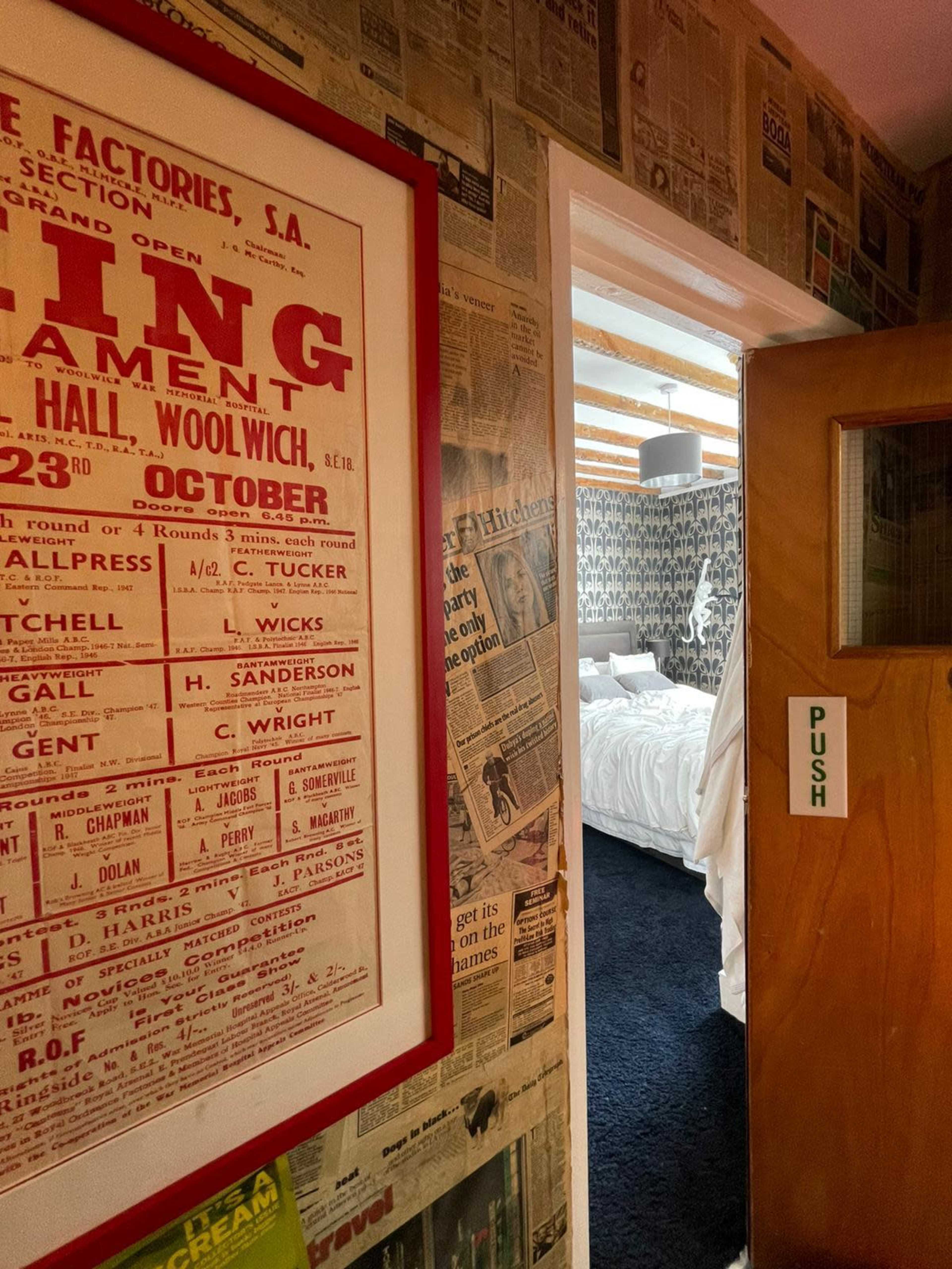 The image shows a hallway with a wall covered in newspaper clippings, leading to a room with a bed and patterned wallpaper.