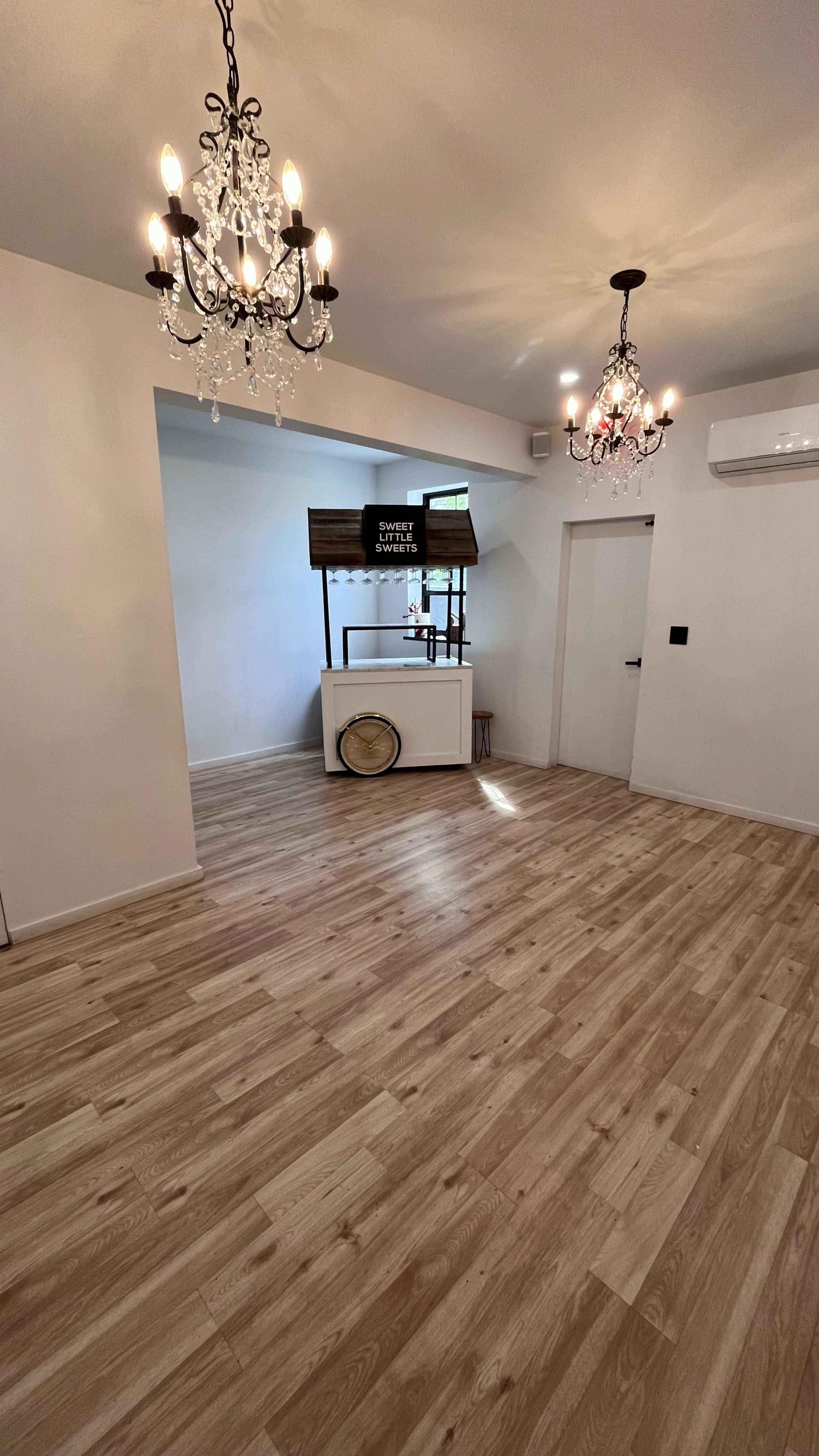 A well-lit, minimalistic interior space featuring wooden flooring, two decorative chandeliers, and a display counter with a television screen against a white wall.
