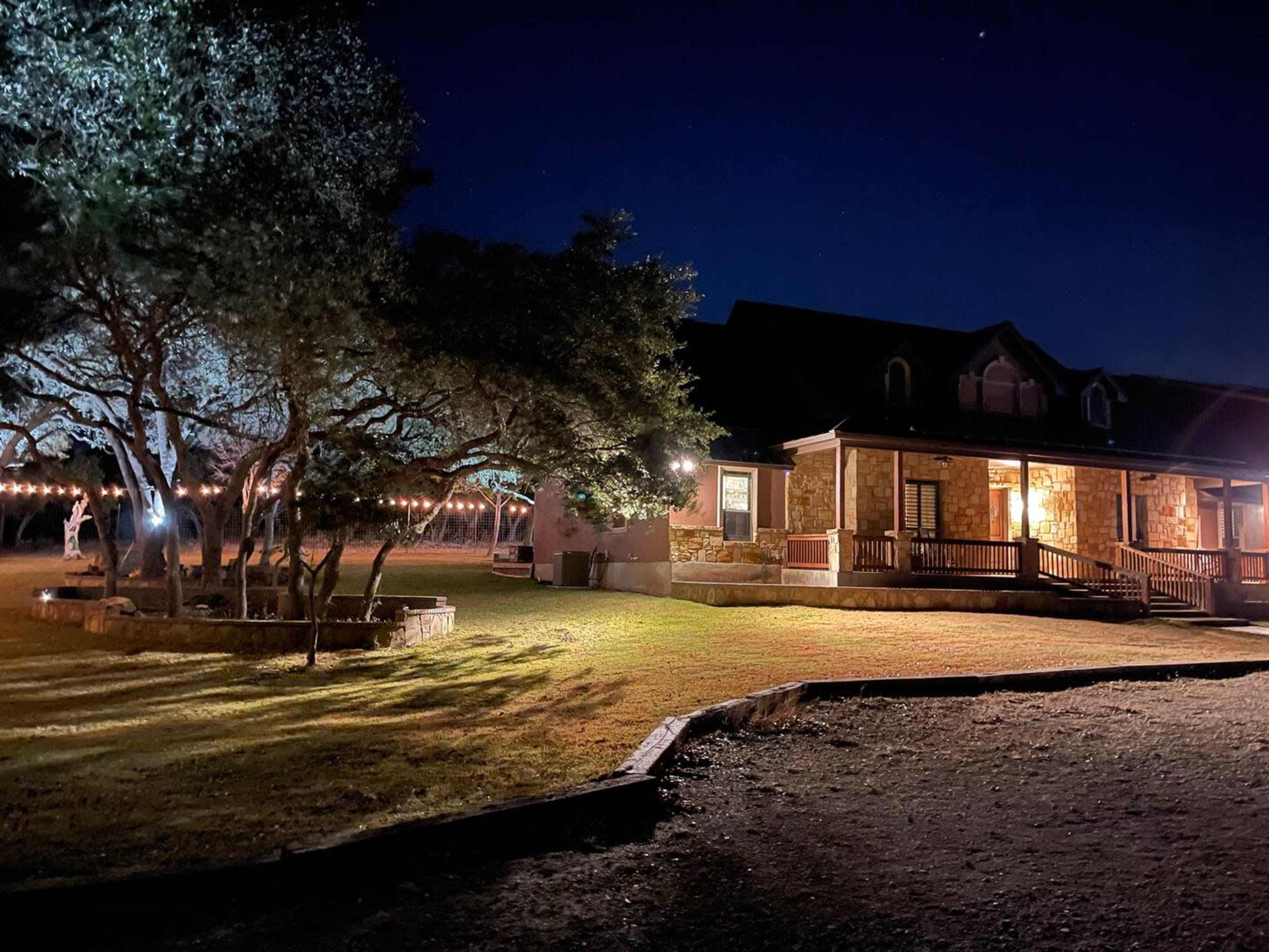 A well-lit stone house is surrounded by trees and a grassy yard at night, with decorative string lights illuminating the area.