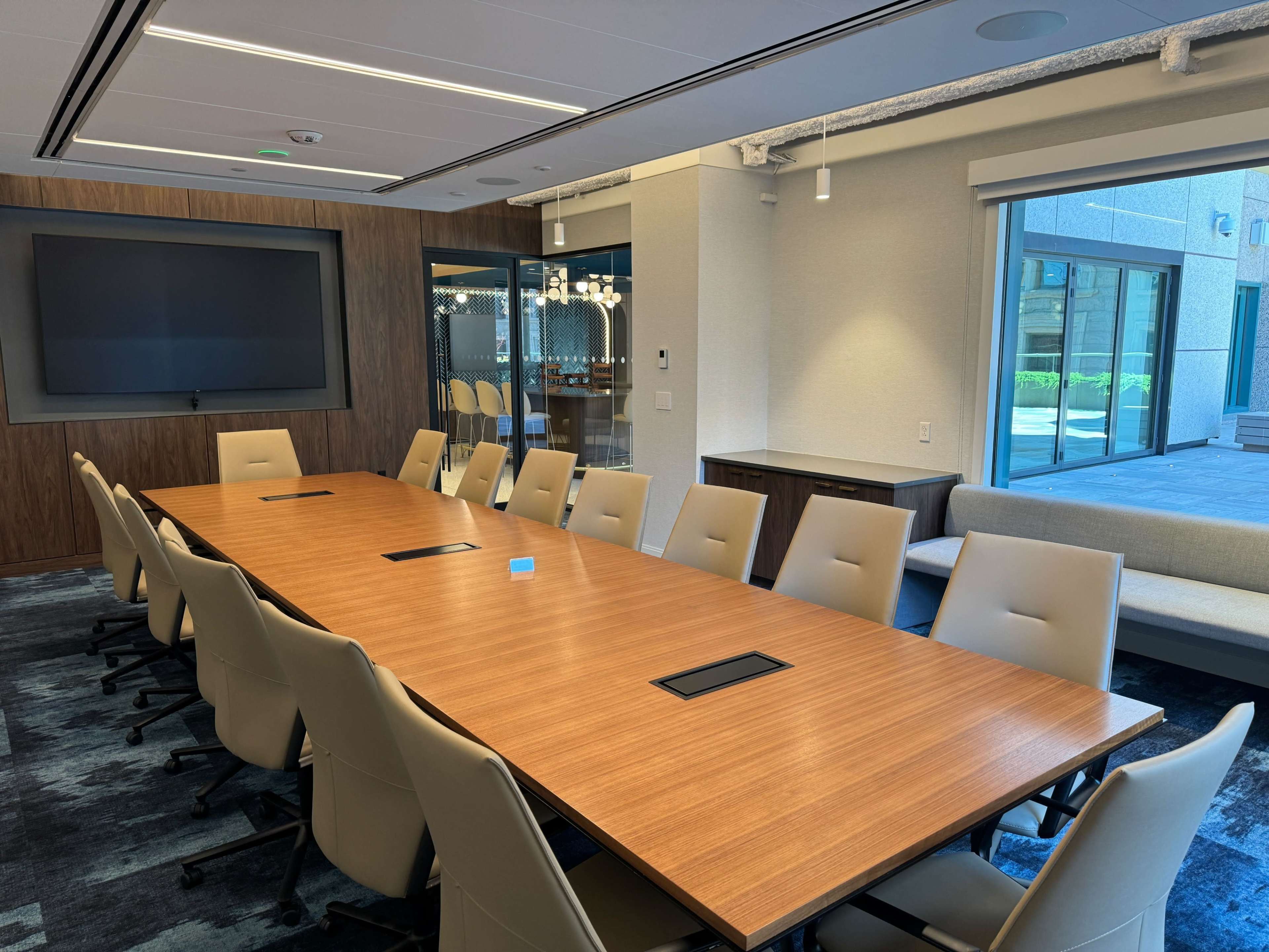 A spacious conference room features a large wooden table surrounded by beige chairs, with a wall-mounted screen and large windows overlooking an outdoor area.