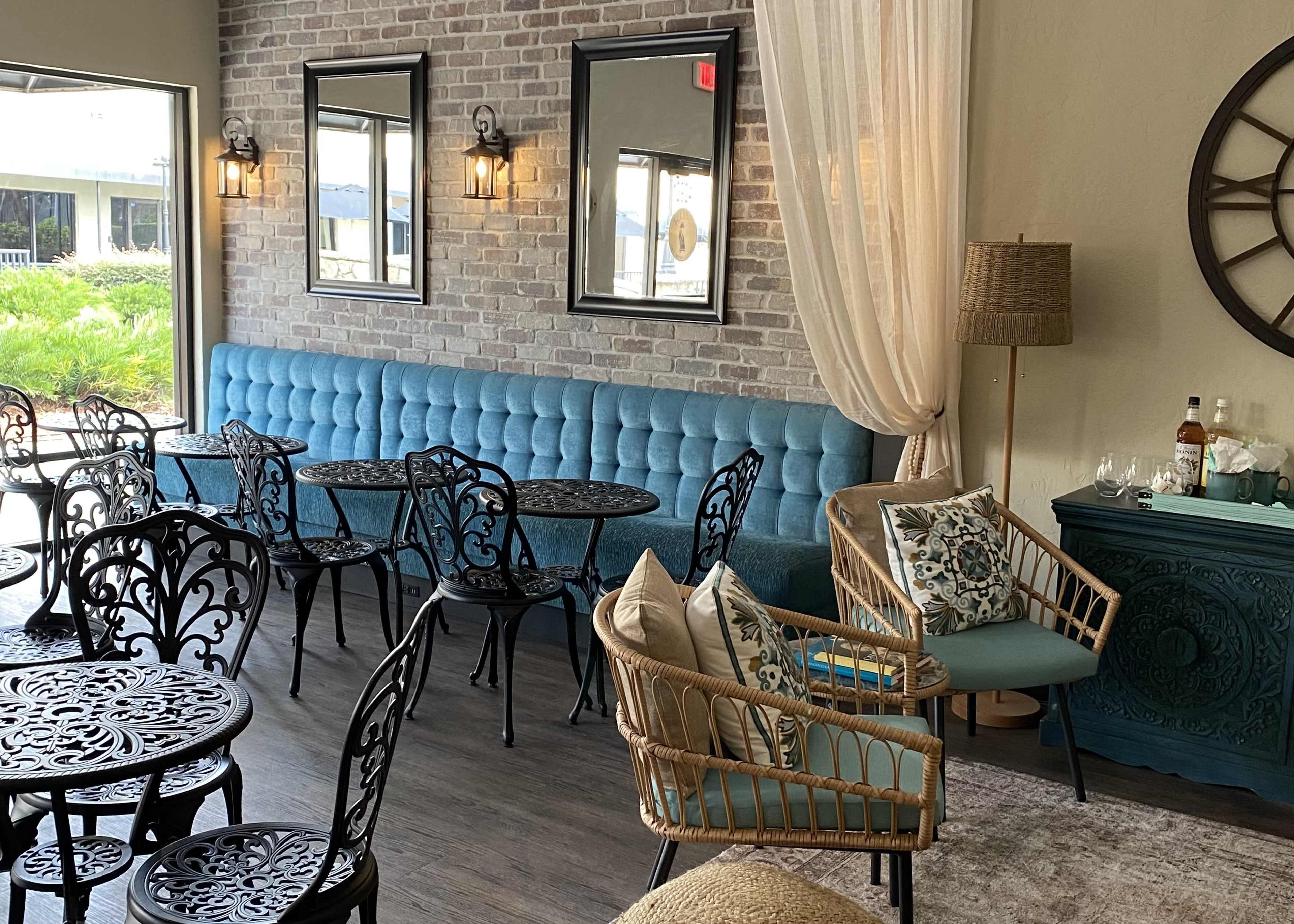 A cozy café interior featuring a blue tufted bench against a brick wall, black wrought iron tables and chairs, and a small seating area with rattan furniture.