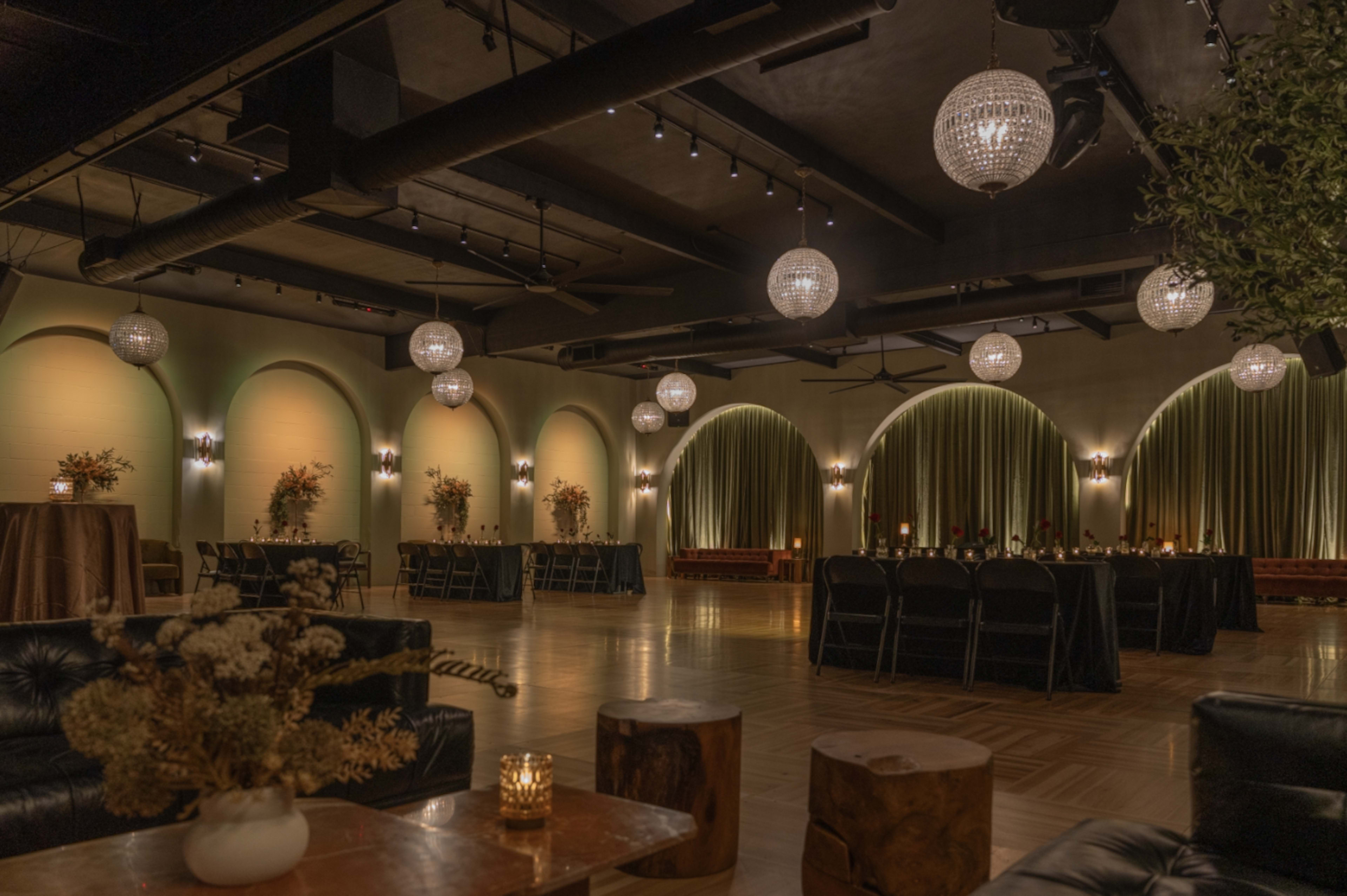 An elegant event space with arched walls, dim lighting, and round chandeliers, featuring tables and seating arranged for an occasion.