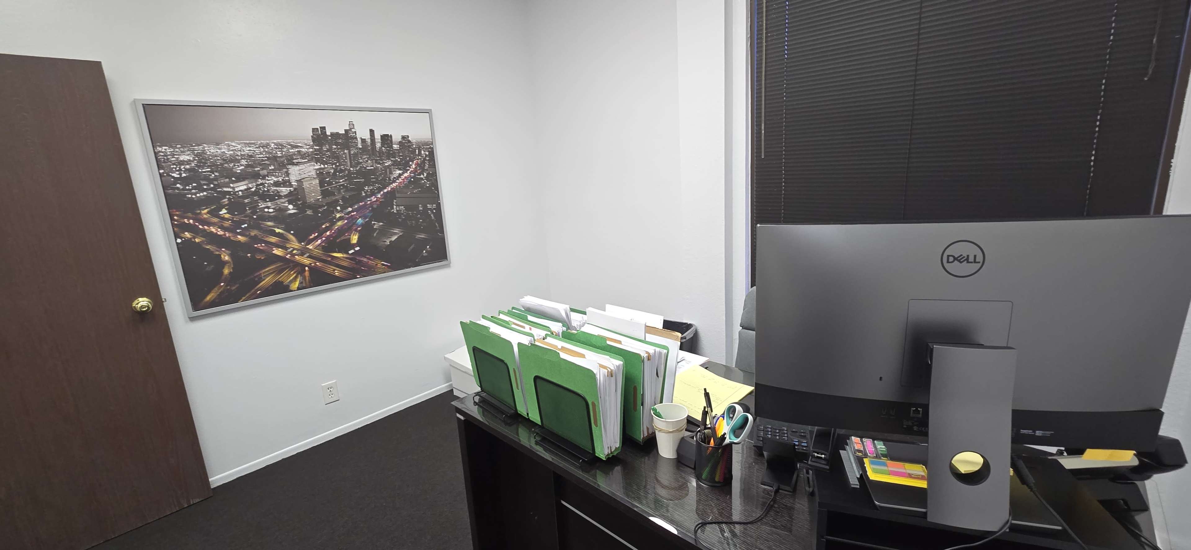 A corner of an office featuring a desk with a computer, stacks of green file folders, and a wall-mounted black-and-white city skyline photograph.