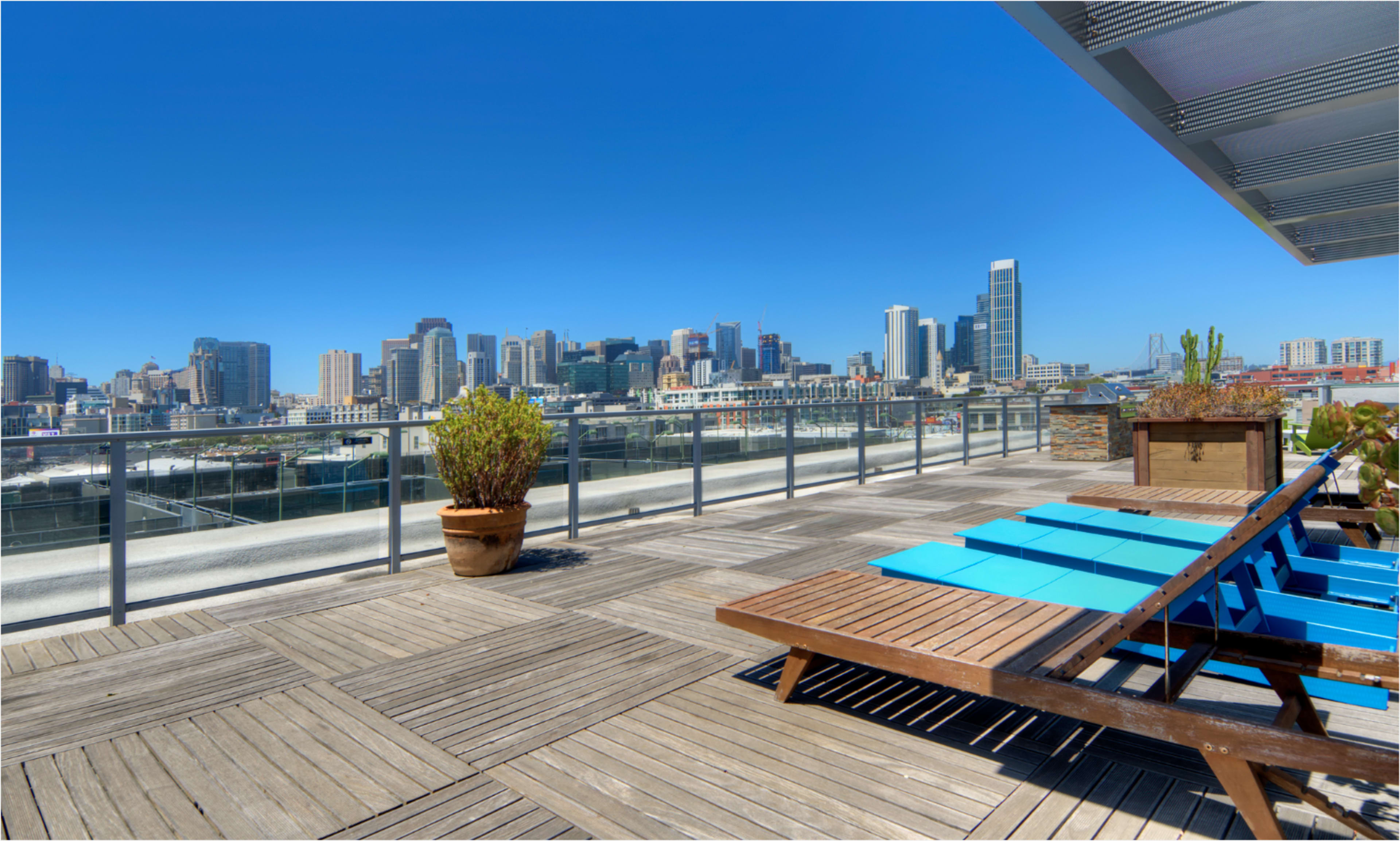 The image shows a rooftop terrace with wooden decking and blue lounge chairs, overlooking a city skyline under a clear blue sky.