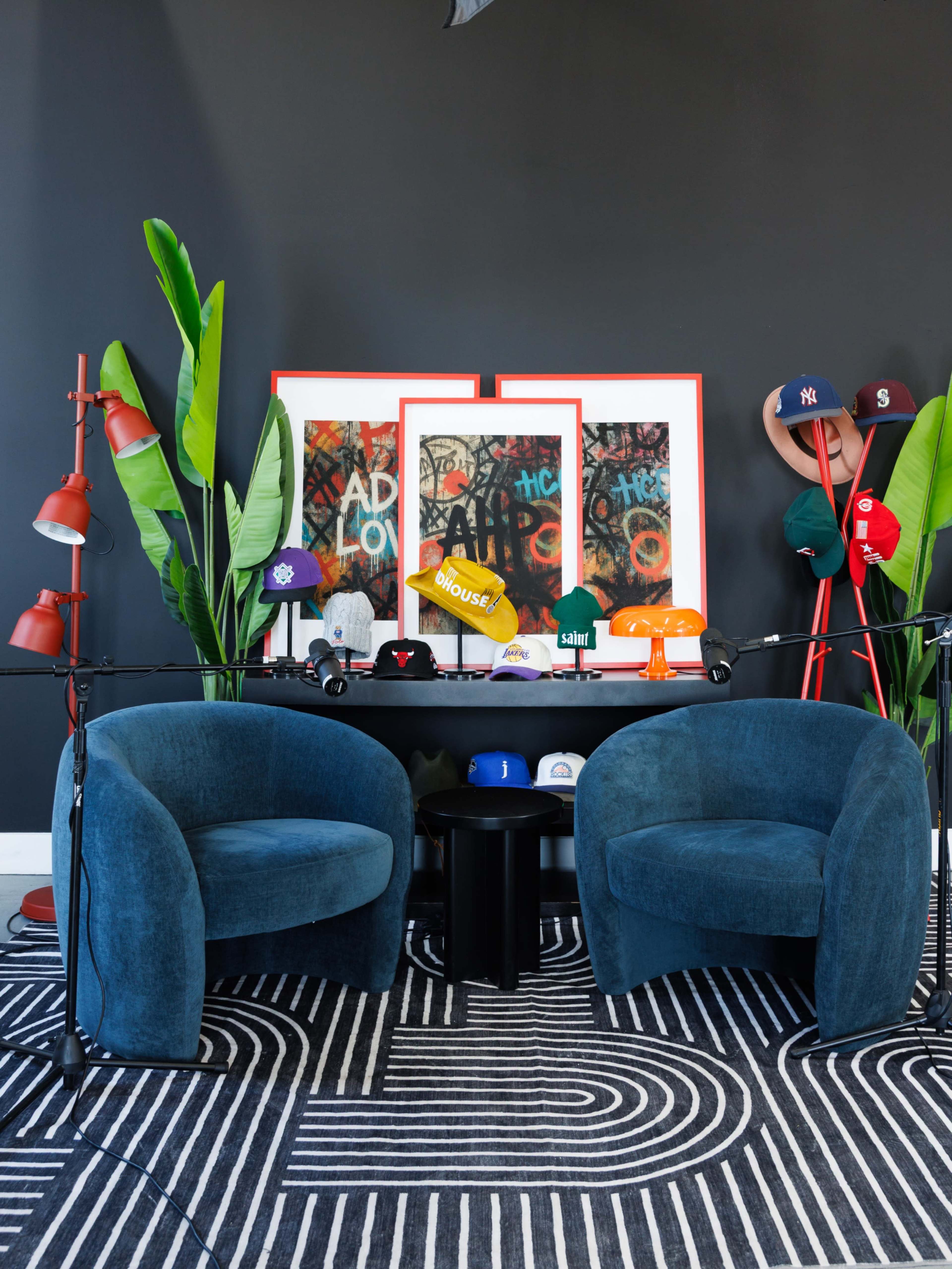 The image features two blue lounge chairs facing a black wall adorned with framed artwork and various caps displayed on a table and hooks.