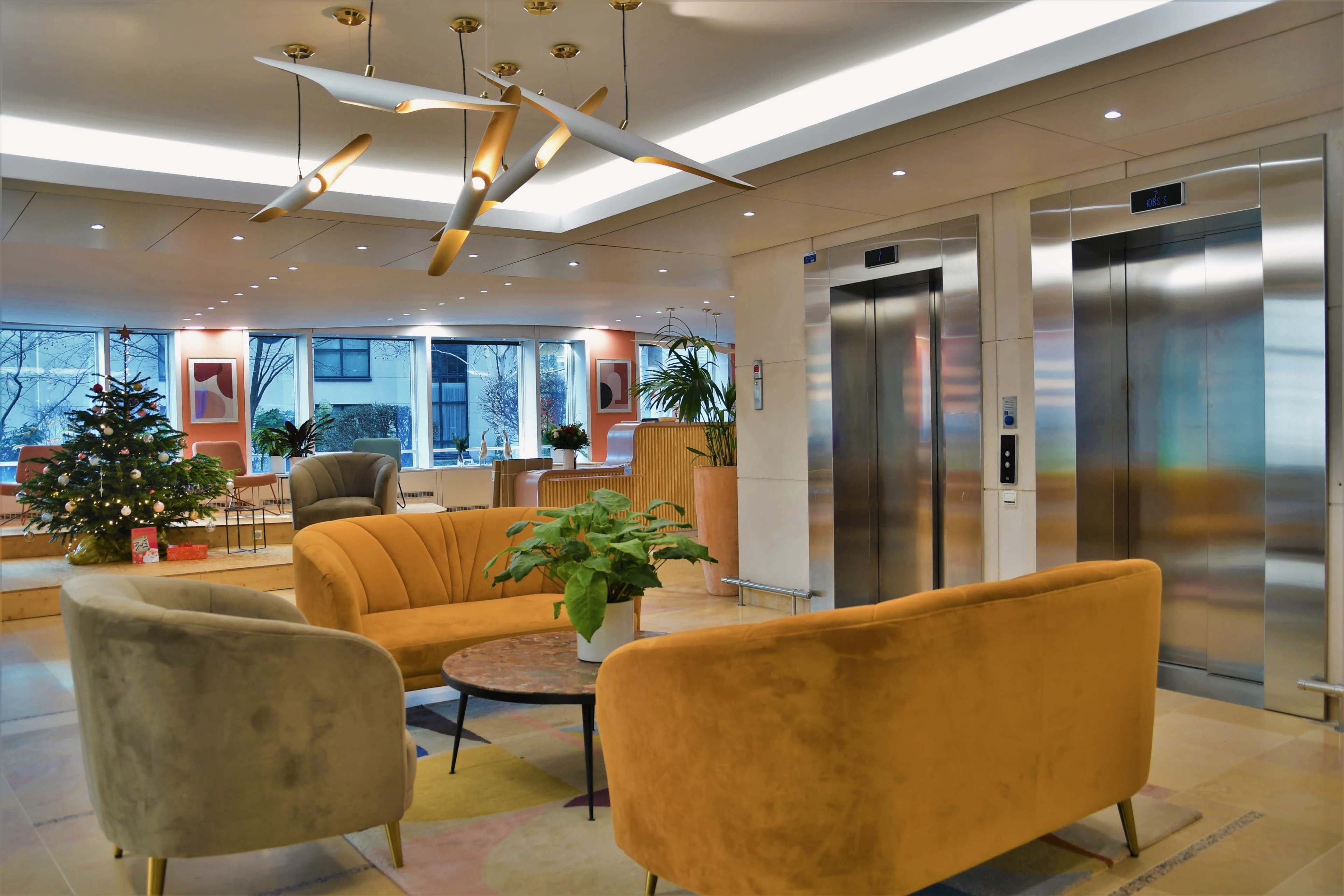 A modern lobby features two upholstered sofas, a circular coffee table, and a decorative tree, with glass elevators visible in the background.