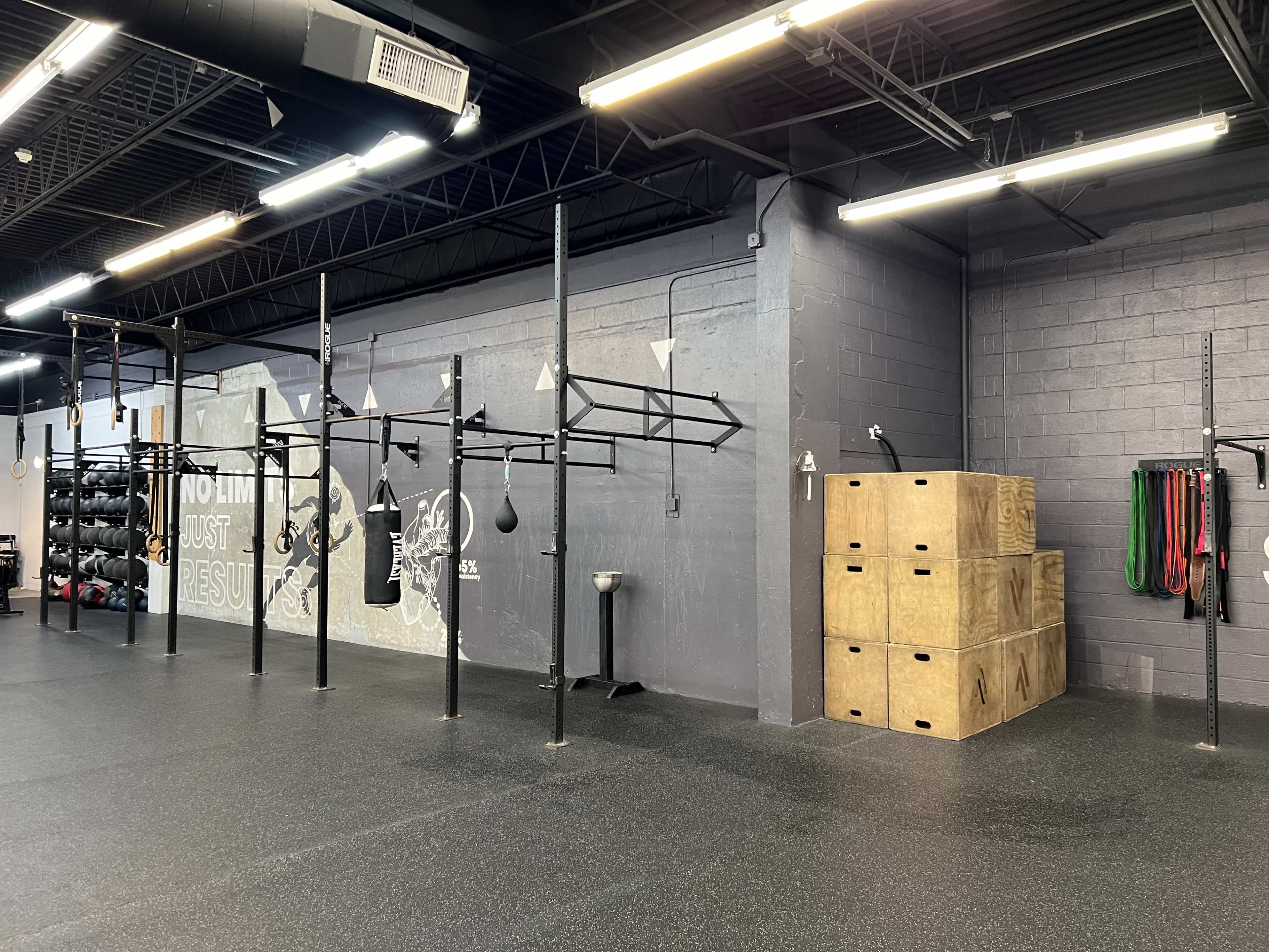 The image shows a fitness gym with wall-mounted pull-up bars, boxing bags, and stacked wooden boxes against a gray wall.