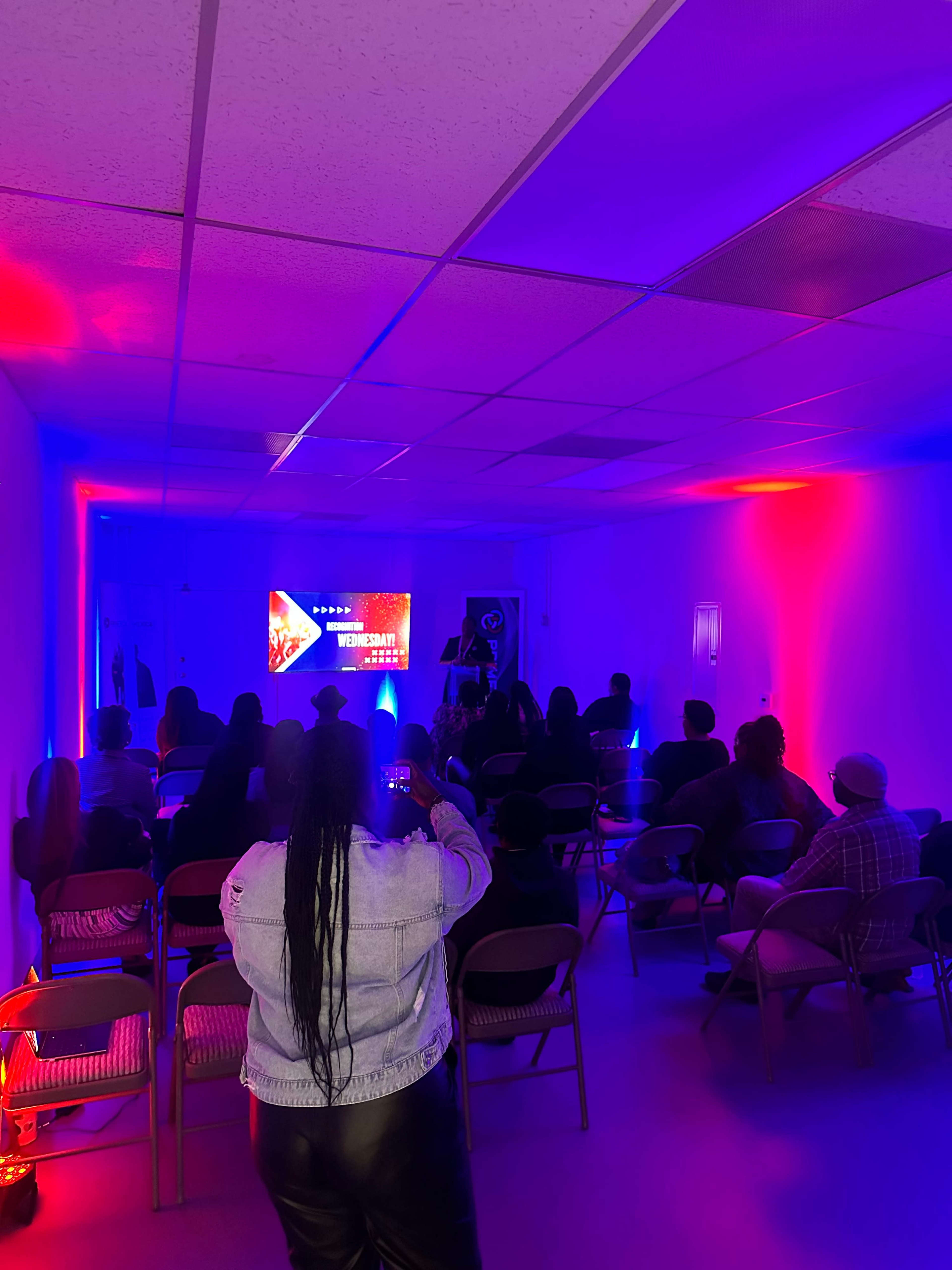 A speaker stands at a podium in a room illuminated with blue and red lights, addressing an audience seated in chairs facing a large screen displaying an event title.