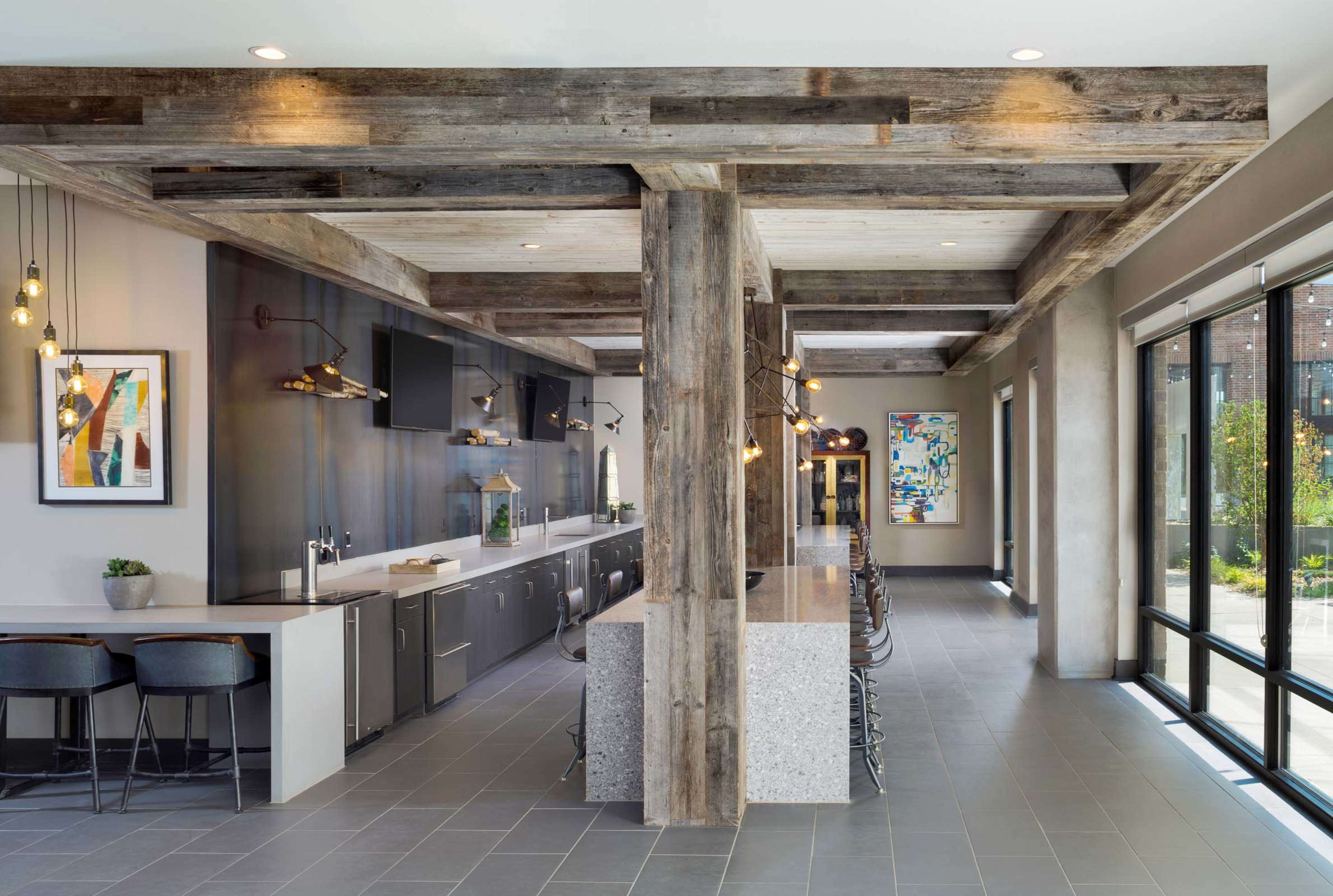 The image shows a modern kitchen and dining area with a long, sleek counter, industrial-style lighting, and large windows overlooking an outdoor space.