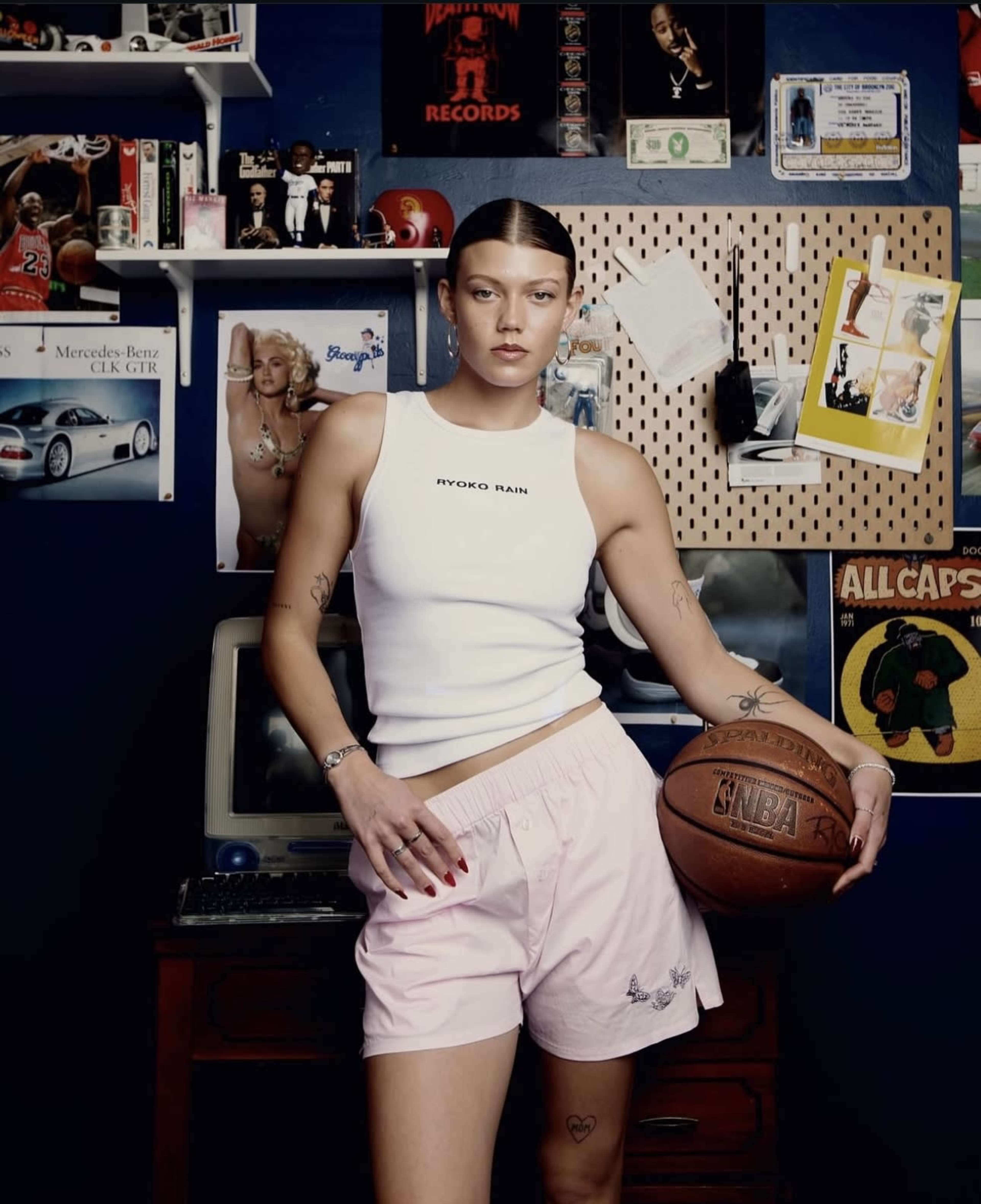 A person stands in front of a wall adorned with various posters and memorabilia, holding a basketball while wearing a white tank top and pink shorts.