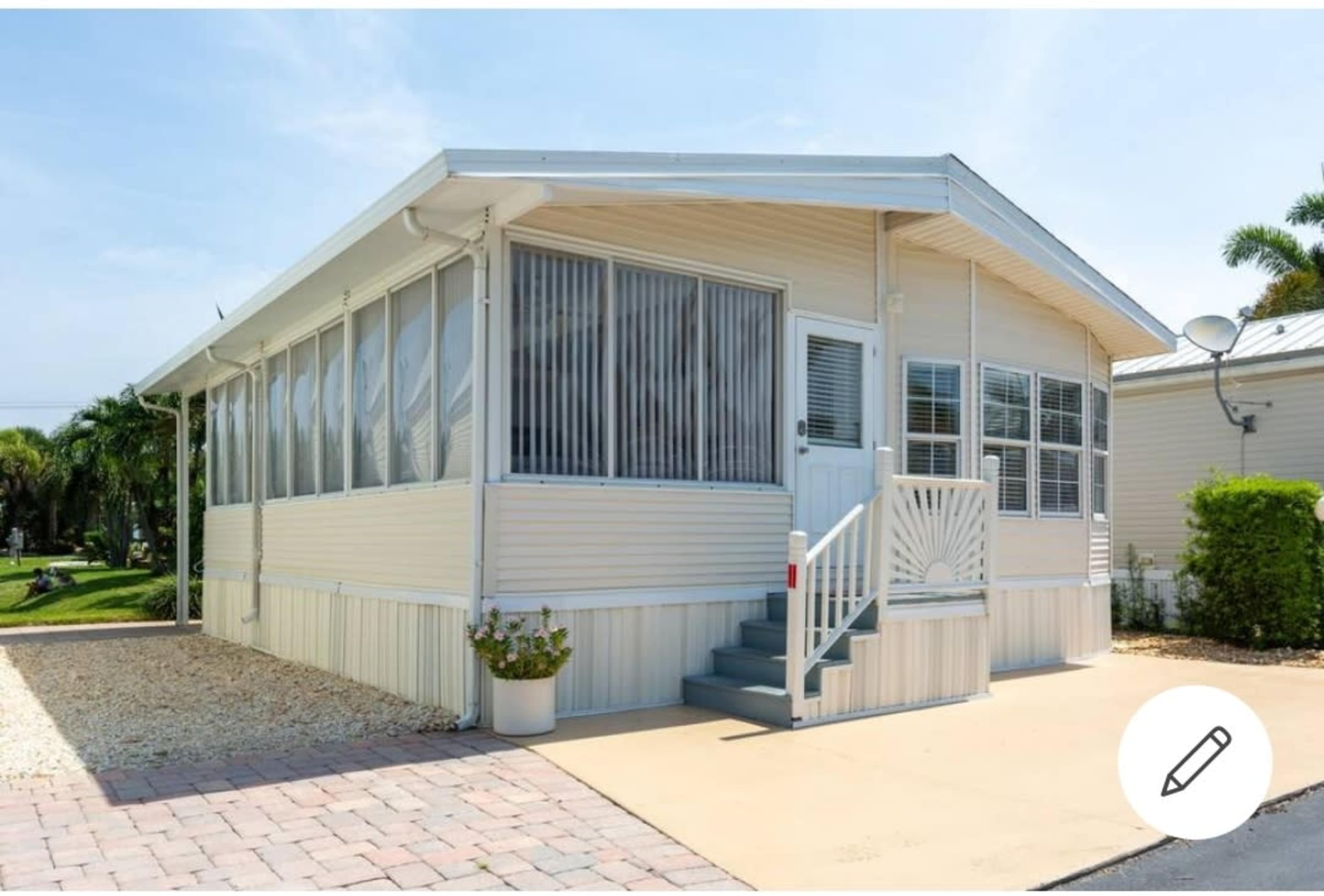 A single-story manufactured home with a front porch, surrounded by a stone pathway and landscaped greenery.