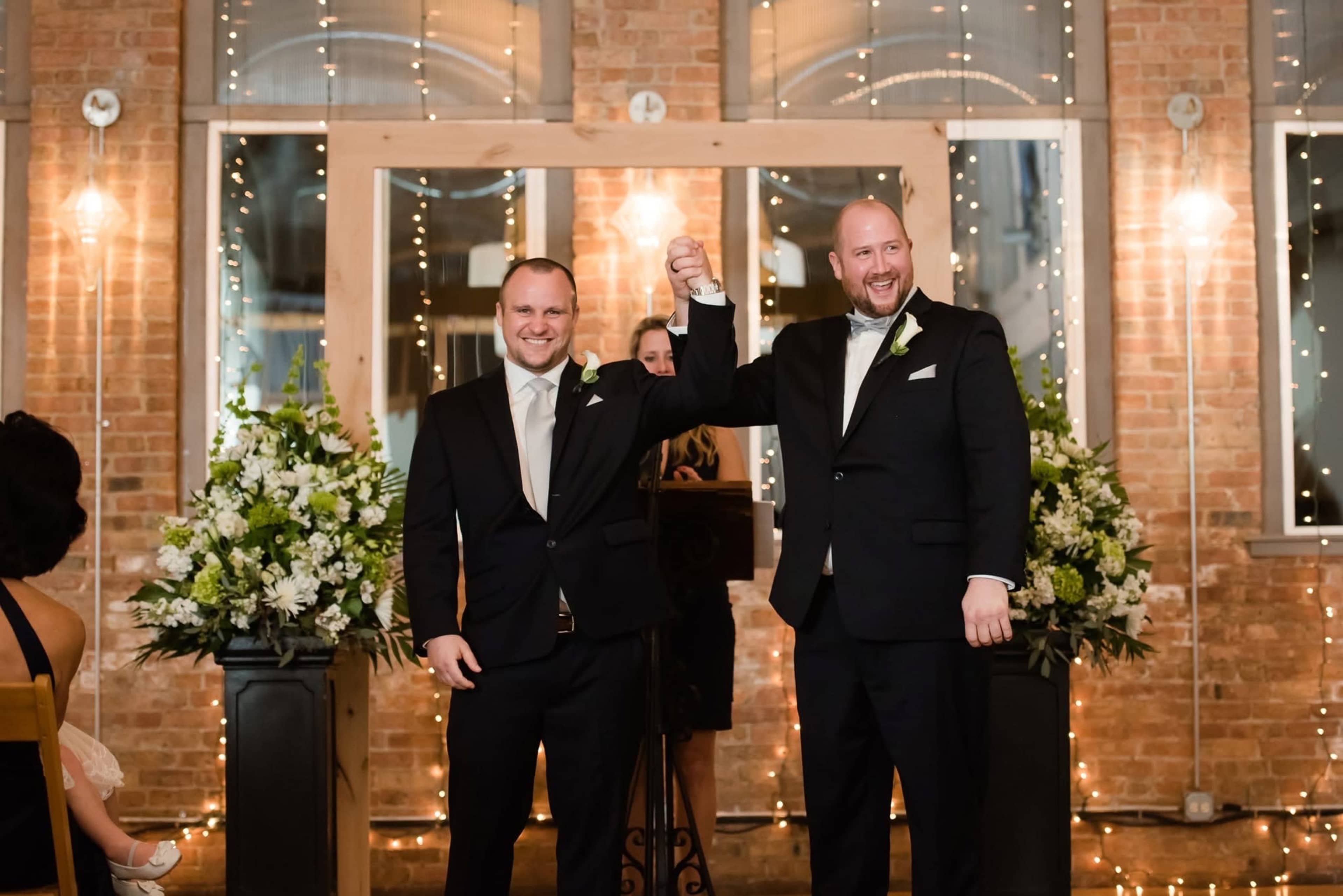 Two men in black tuxedos raise their arms in celebration during a wedding ceremony, surrounded by floral arrangements and twinkling lights on a brick wall.
