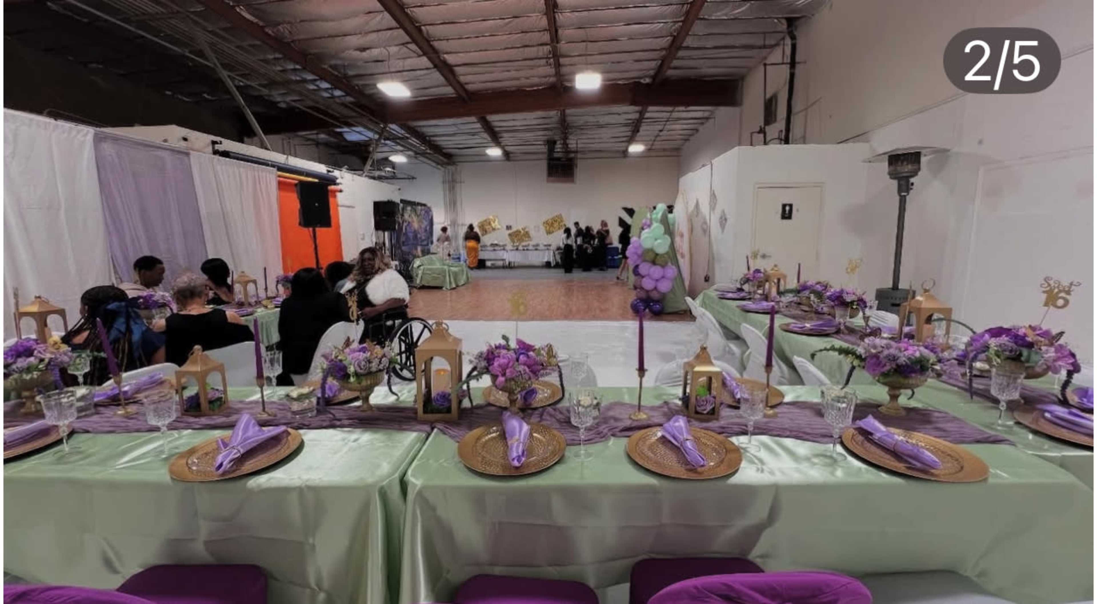 A decorated banquet space features tables set for a celebration with floral arrangements, purple accents, and a stage area in the background.