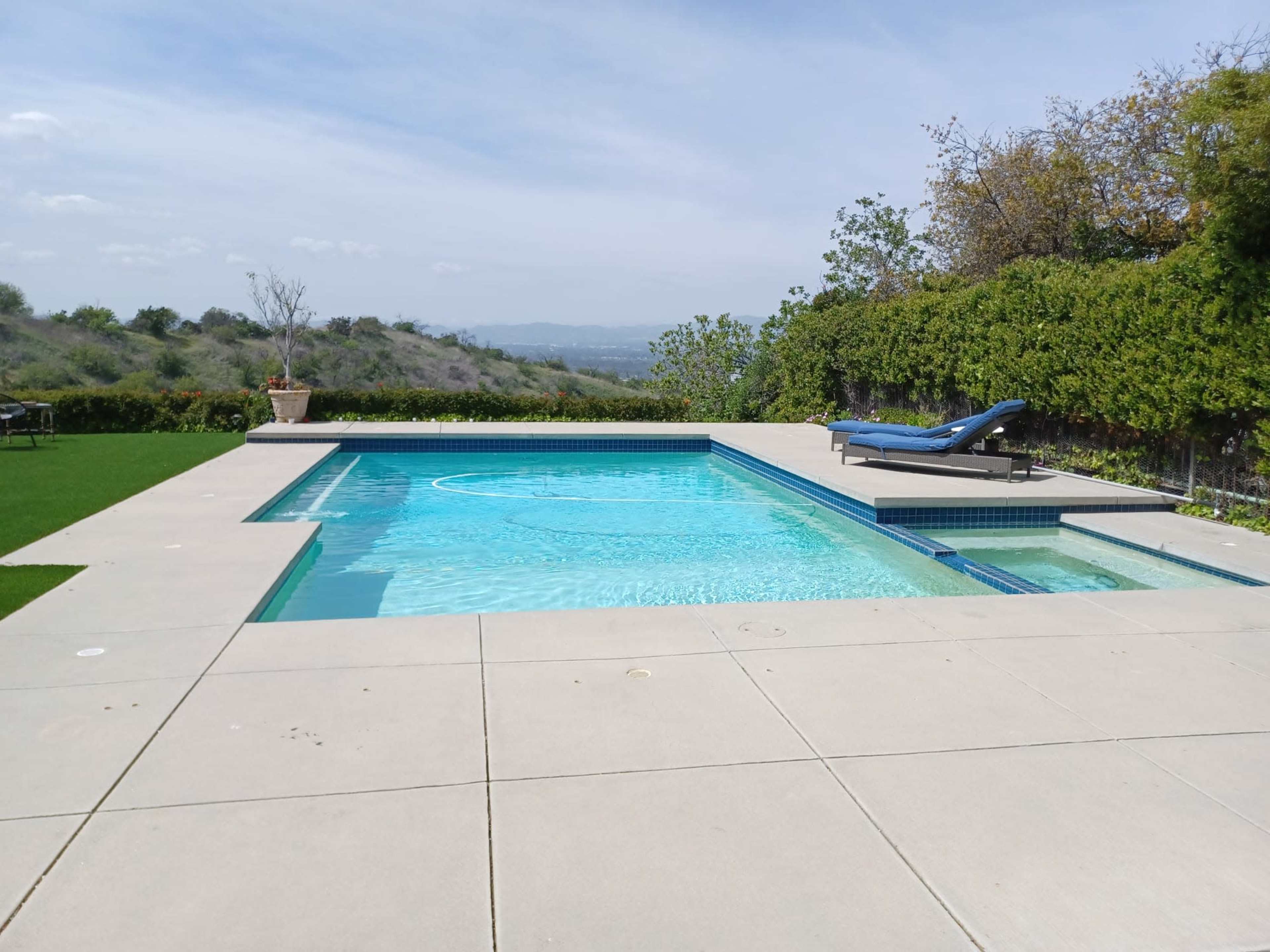 A rectangular swimming pool surrounded by a concrete patio, with a lounge chair positioned on one side.