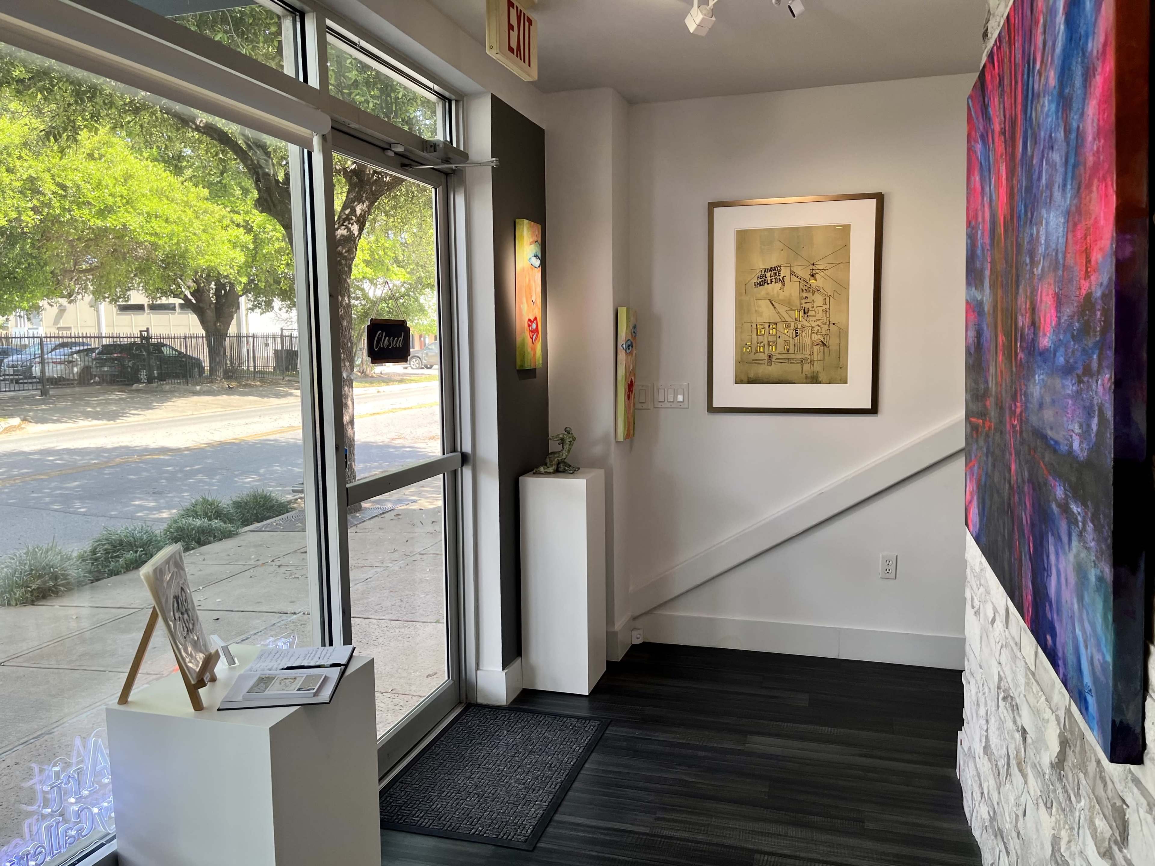 The interior of an art gallery with a view of the entrance, featuring a framed artwork on the wall and a small display table near the door.