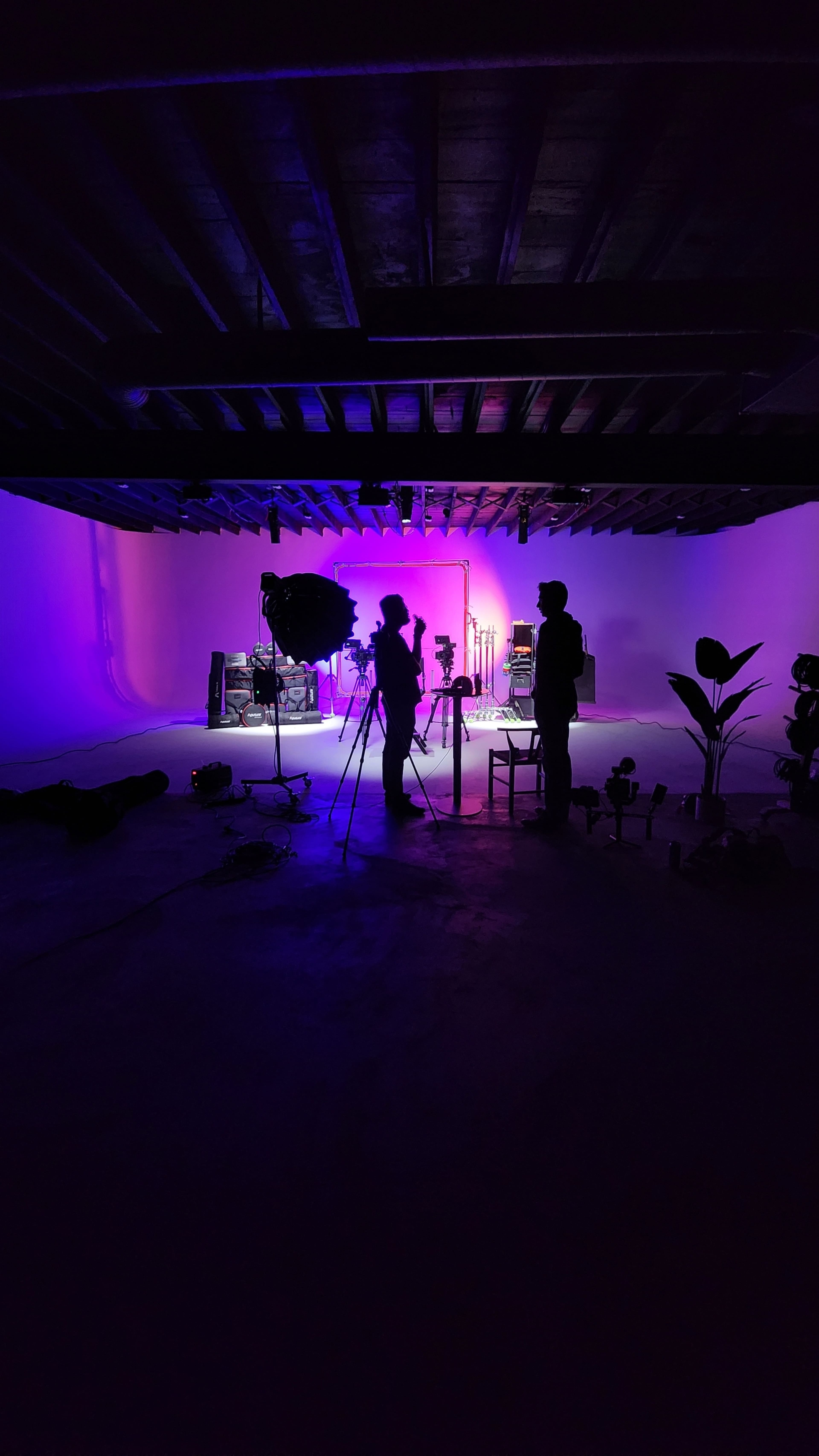 The image shows silhouettes of two people in a dimly lit studio with purple and blue lighting, surrounded by camera equipment and various props.