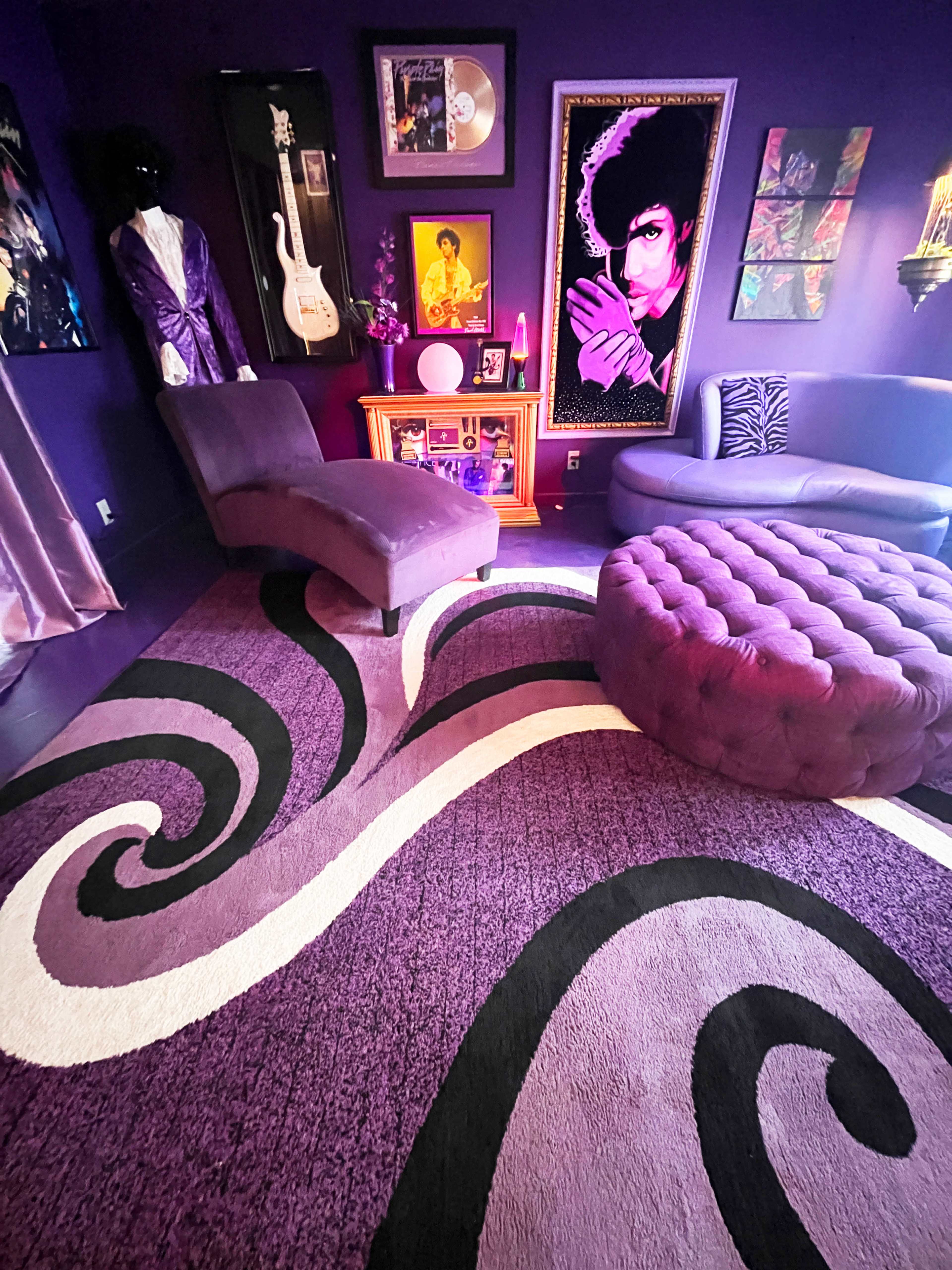The image shows a purple-themed room featuring a curvy rug, a chaise lounge, a round tufted ottoman, and various artwork, including a portrait of a musician.